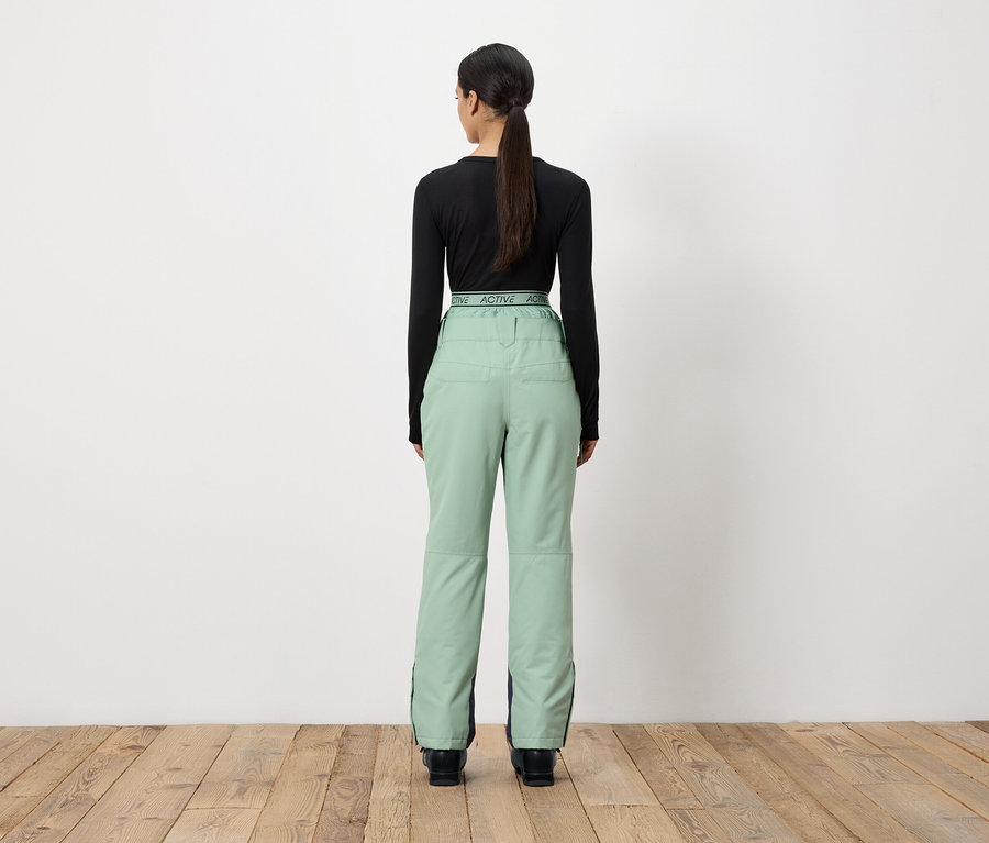 Kobieta tyłem do kamery ma na sobie czarną bluzkę z długim rękawem i jasnozielone spodnie.