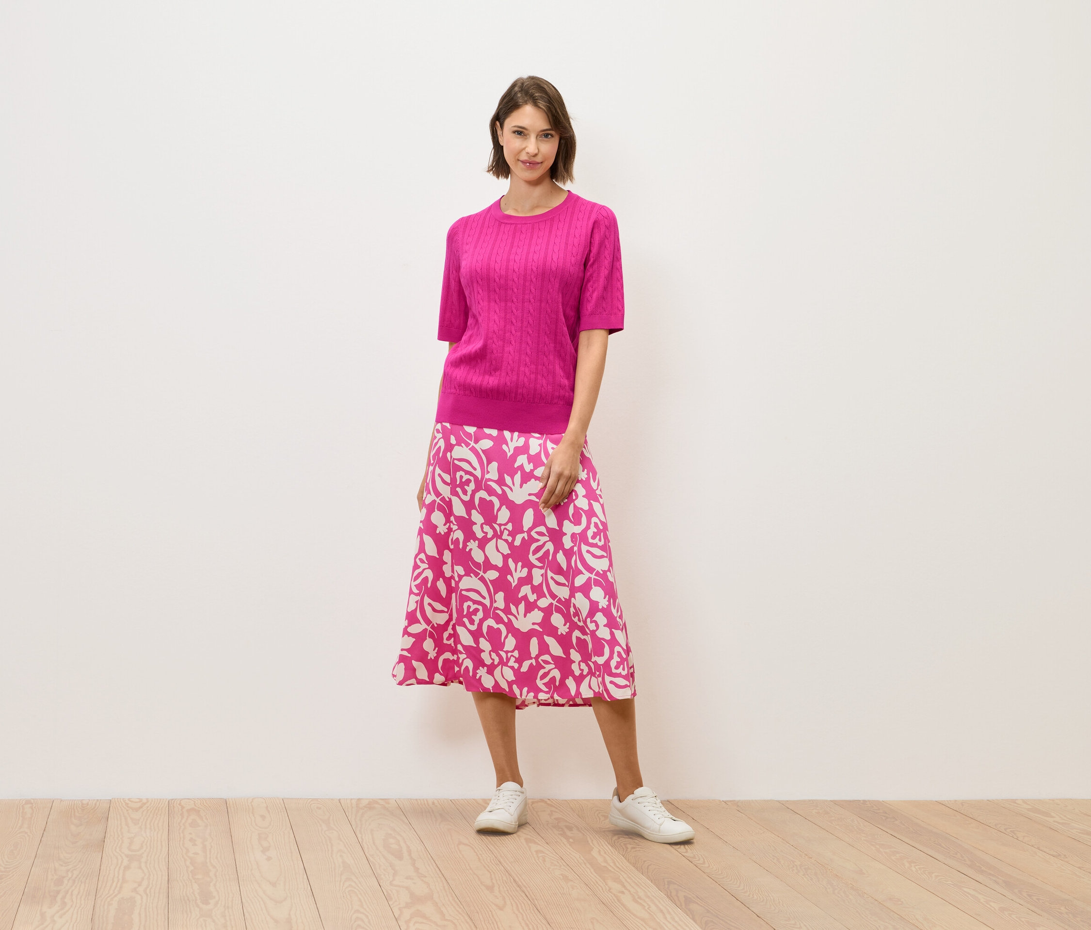 Kobieta w różowym swetrze i spódnicy w kwiaty stoi na drewnianej podłodze.