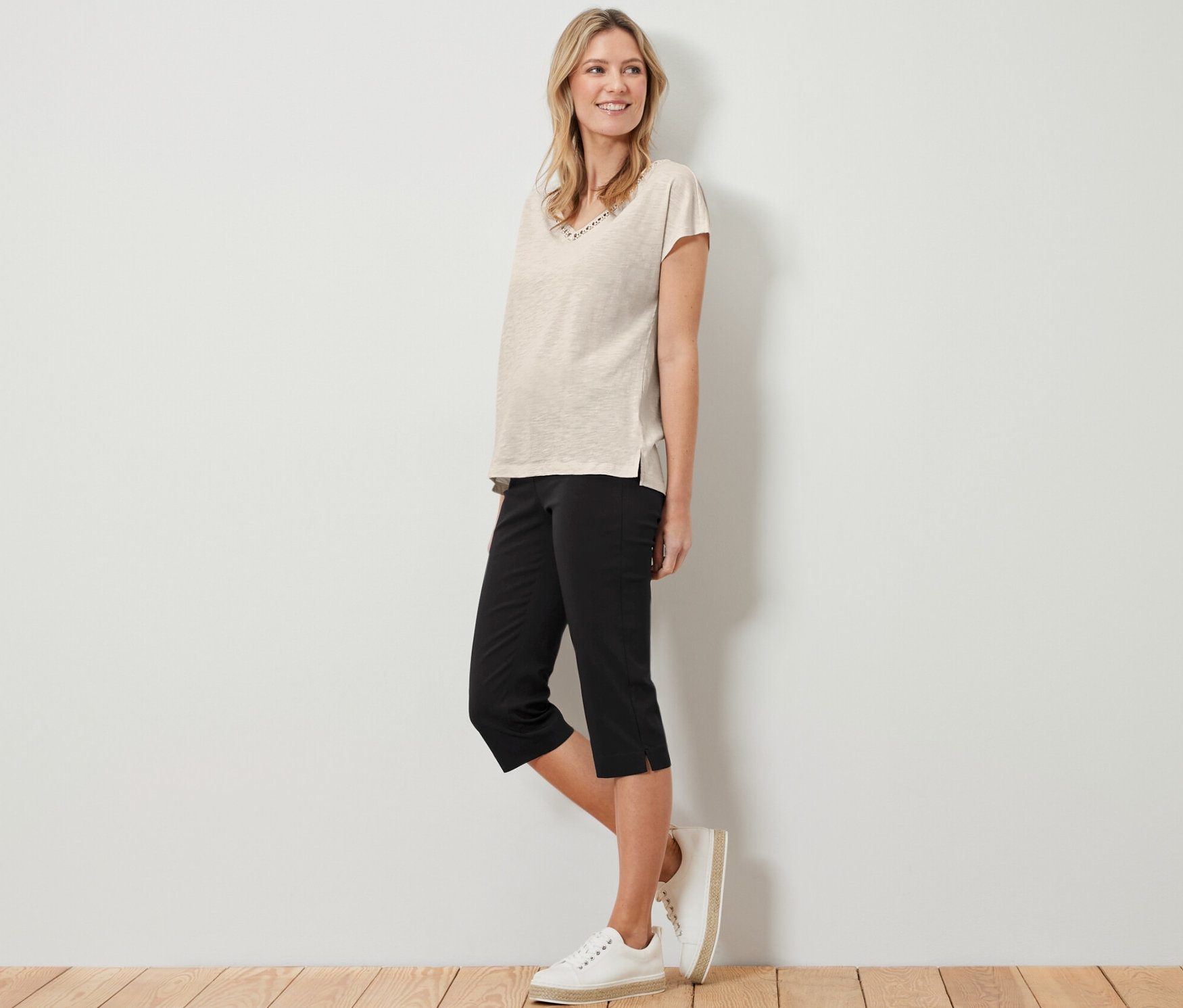 Kobieta stoi, ma na sobie beżowy t-shirt i czarne spodnie stretchowe o długości 3/4.