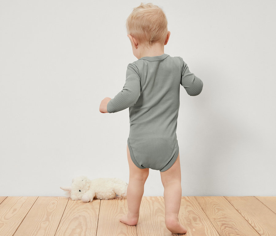 Małe dziecko w body z długim rękawem stoi na drewnianej podłodze obok pluszowego zwierzątka.