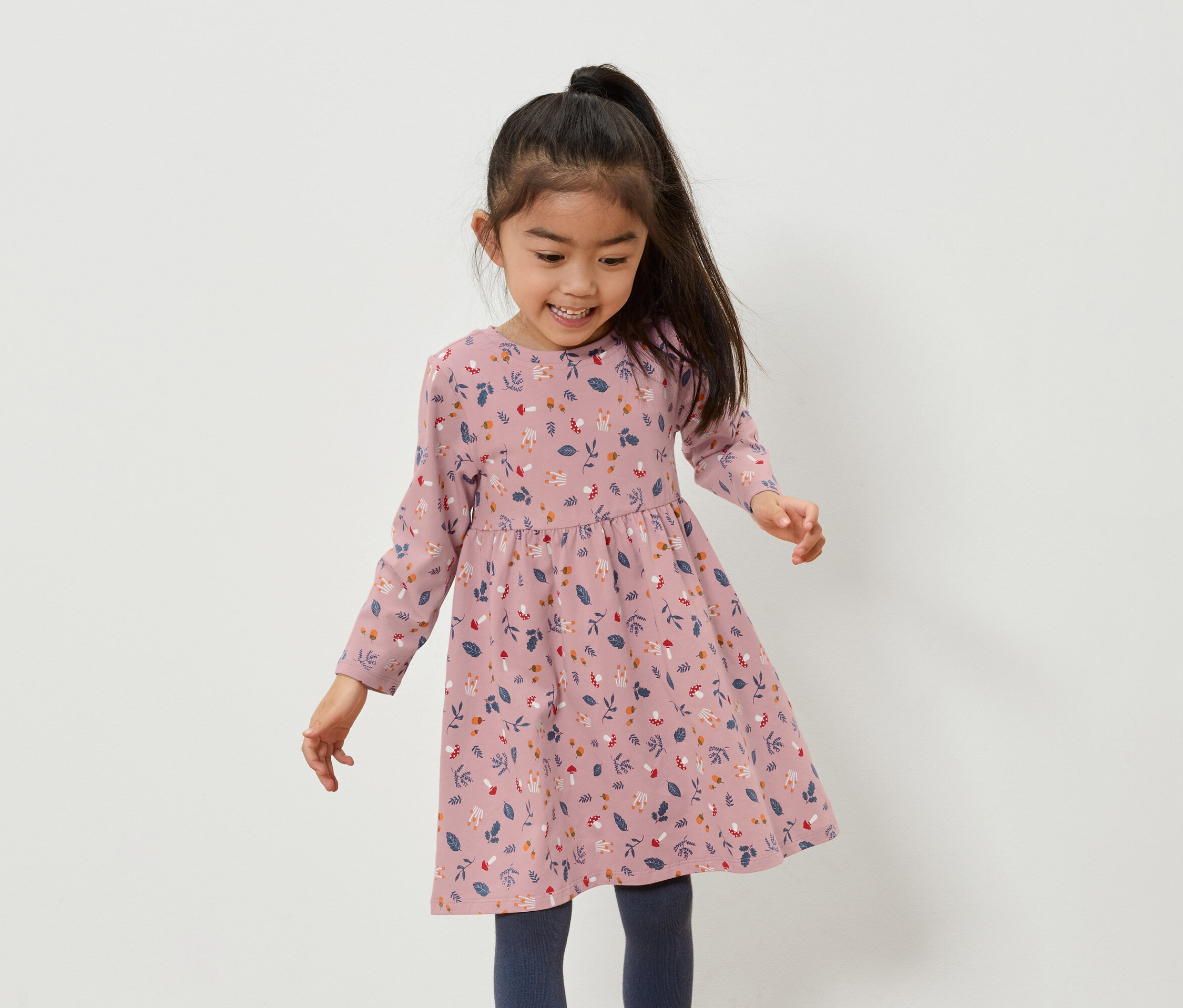 Uśmiechnięta azjatycka dziewczynka w różowej sukience w kwiaty i niebieskich legginsach.