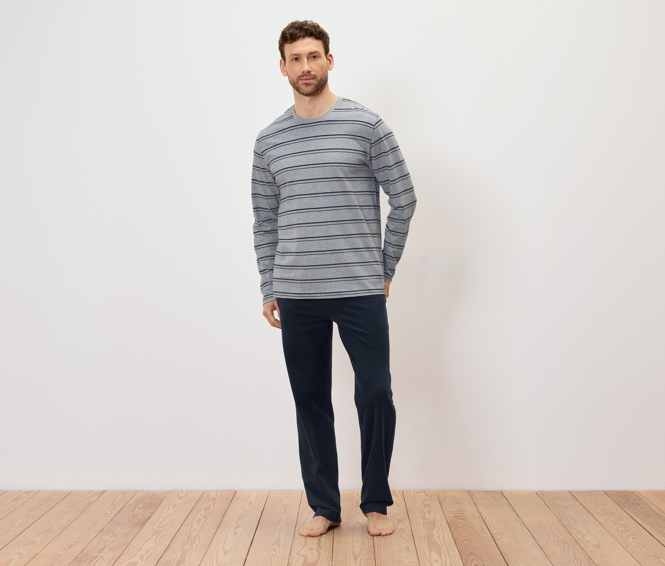 Mężczyzna stoi boso na drewnianej podłodze, ubrany w szarą koszulkę z długim rękawem w paski i granatowe spodnie.