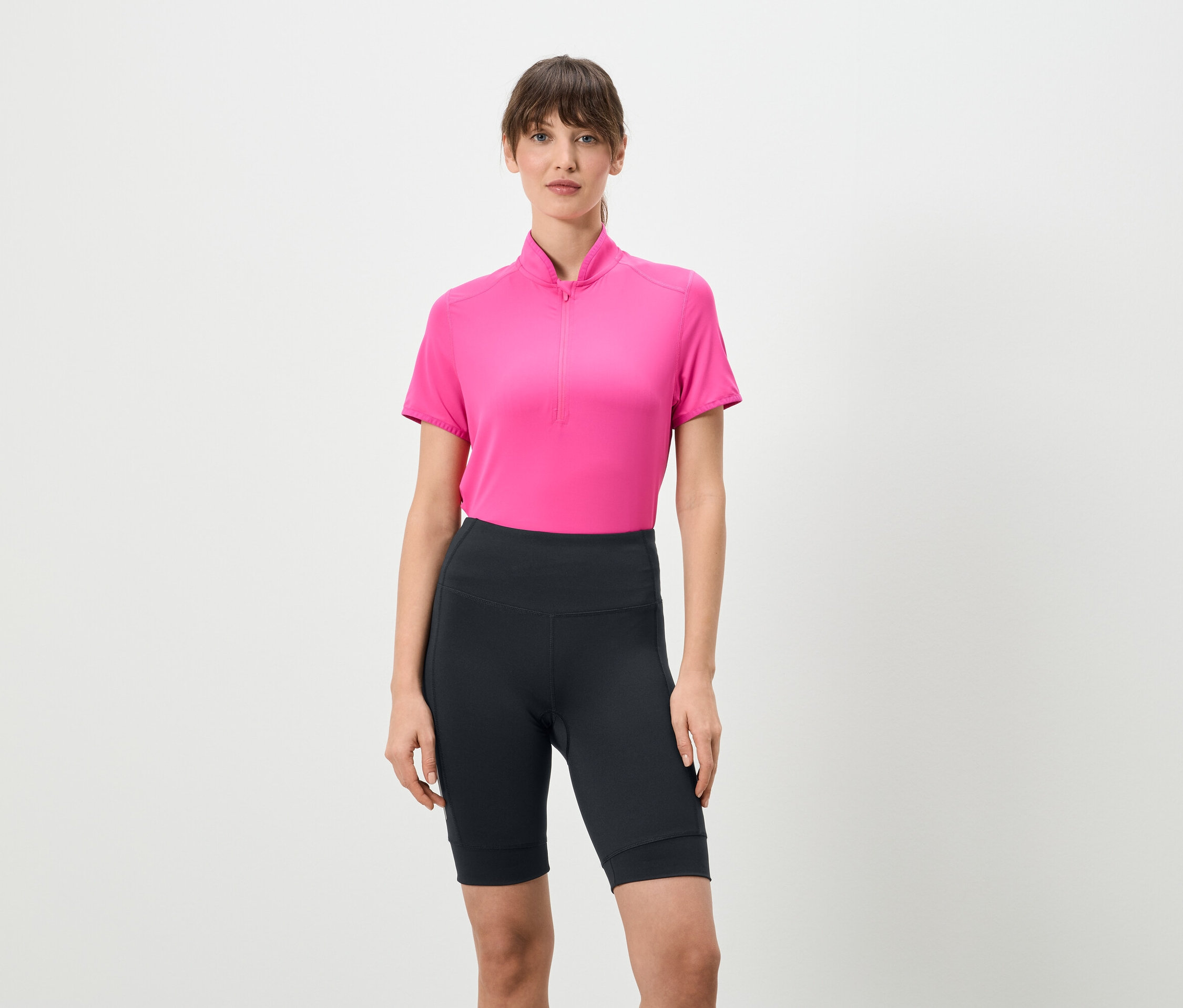 Kobieta stoi na białym tle, ubrana w różową koszulkę z krótkim zamkiem i krótkie, ciemne legginsy.