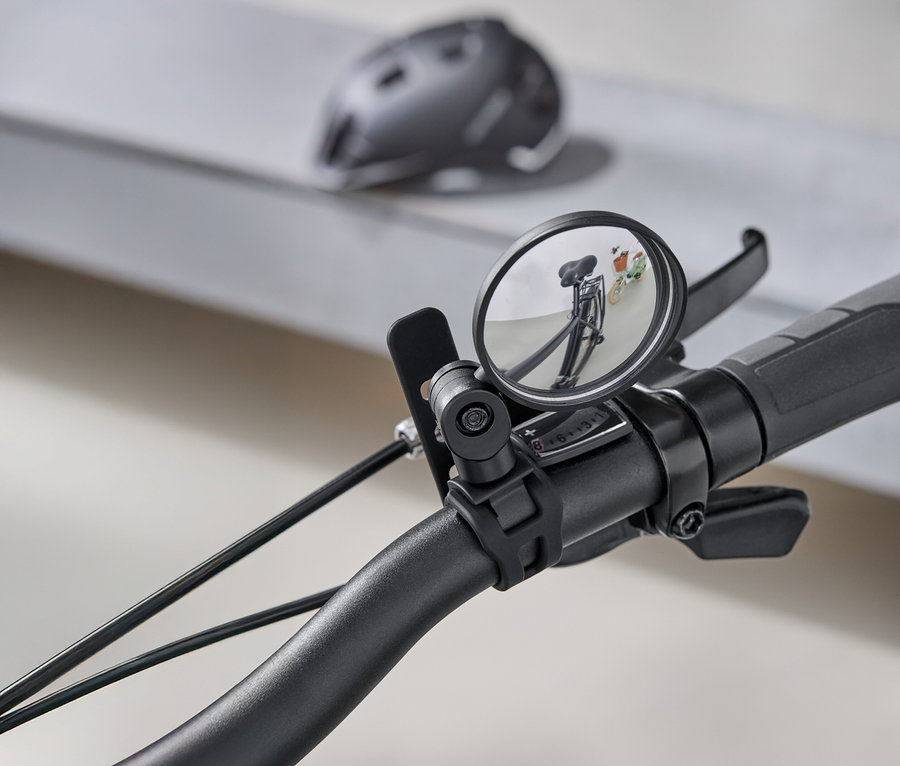 Zbliżenie na kierownicę roweru z okrągłym lusterkiem wstecznym, w którym odbija się droga za rowerem. W tle widać szary kask rowerowy.