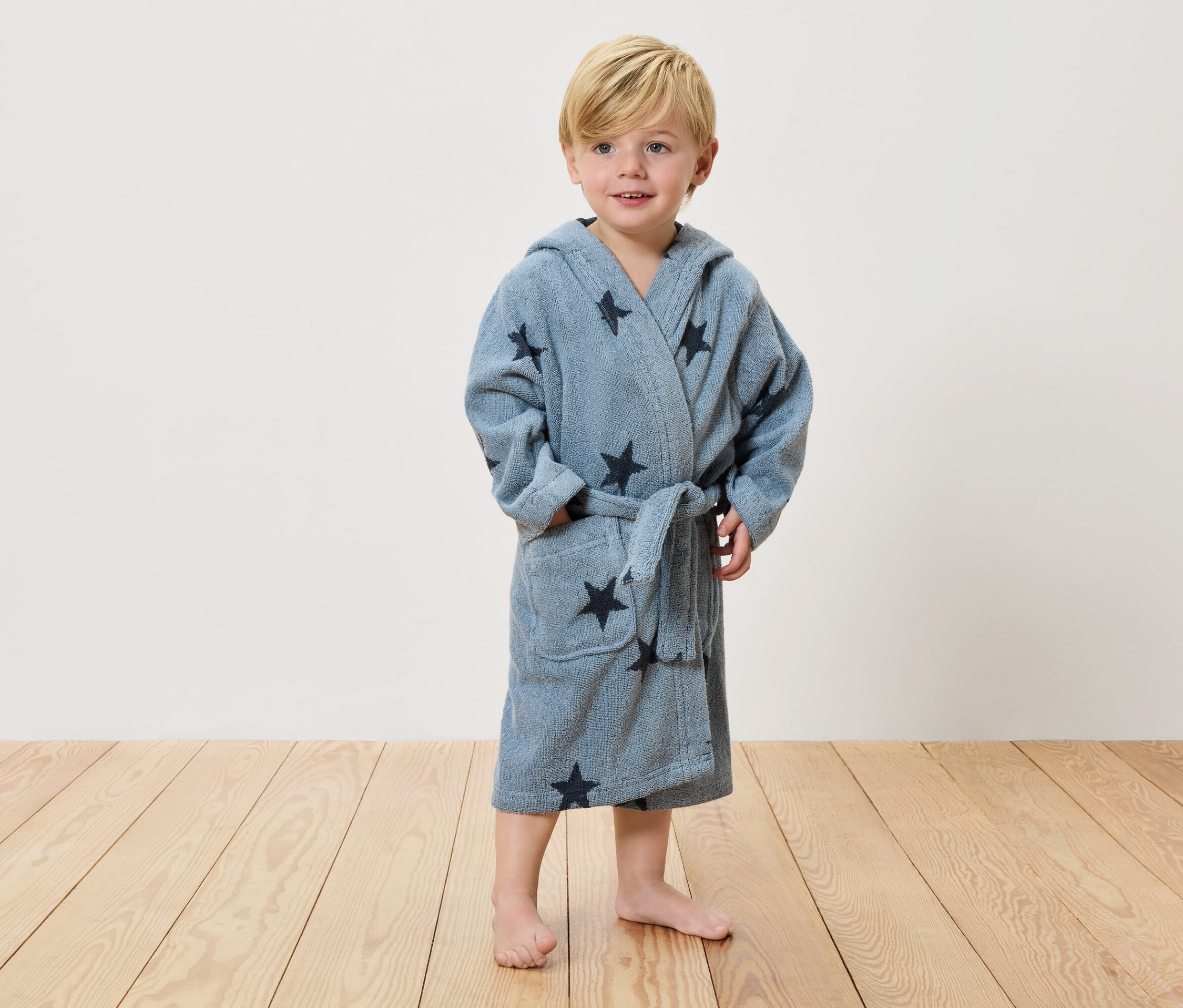 Mały chłopiec z blond włosami stoi boso na drewnianej podłodze, ubrany w jasnoniebieski szlafrok w ciemne gwiazdki.