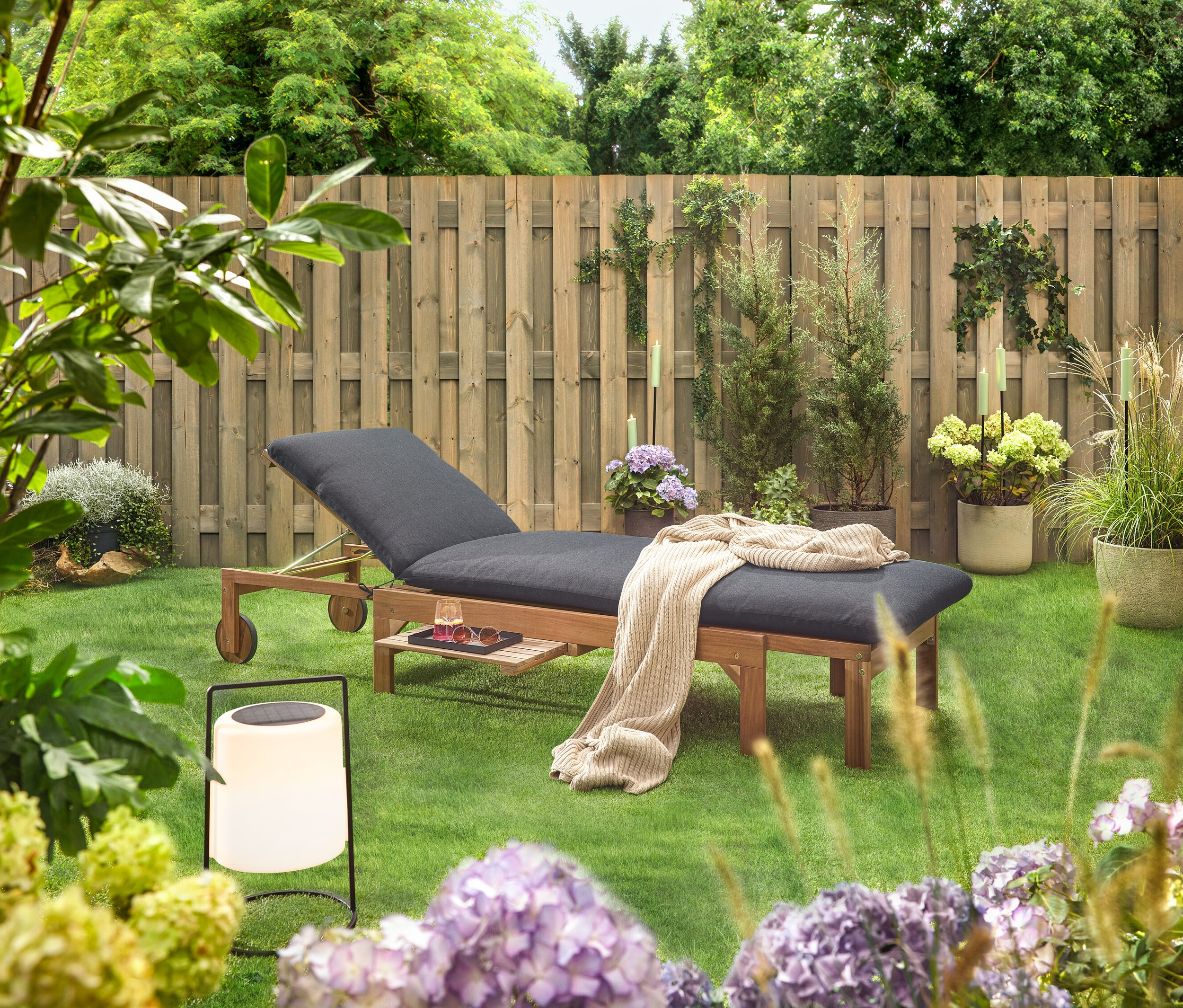 Scena ogrodowa z drewnianym leżakiem z czarną tapicerką, kocem, napojem, okularami przeciwsłonecznymi i lampionem na trawniku przed drewnianym płotem.