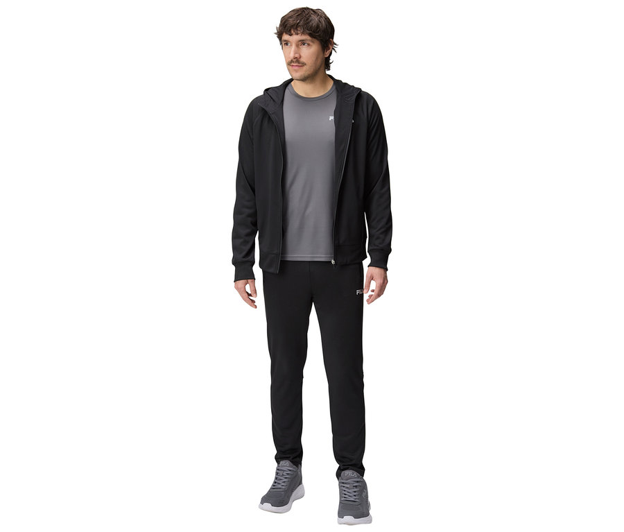 Mężczyzna w czarnej bluzie z kapturem, szarej koszulce i czarnych spodniach dresowych oraz szarych butach sportowych.