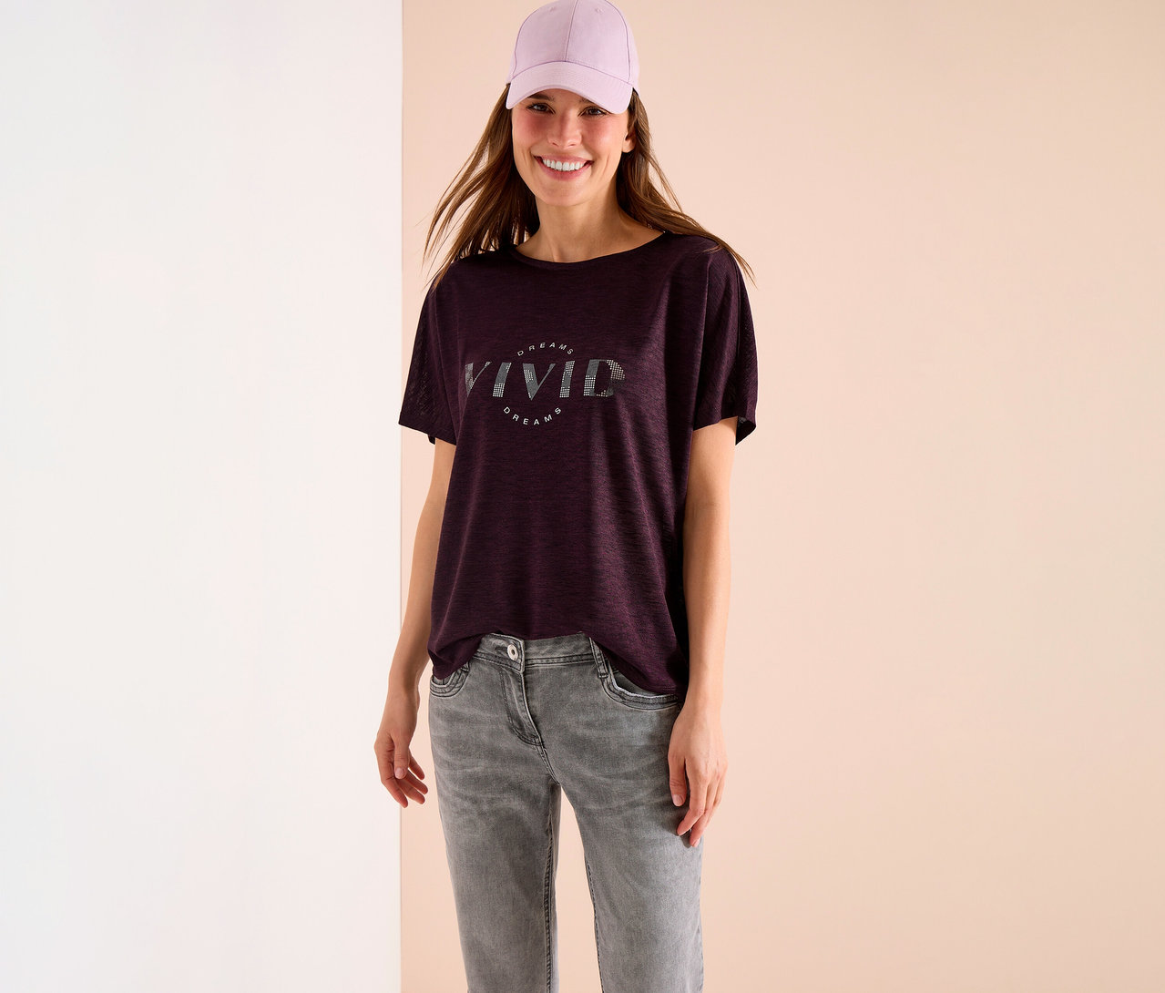 Uśmiechnięta kobieta w fioletowej czapce i bordowym T-shircie z napisem "Vivid Dreams" w szarych dżinsach.