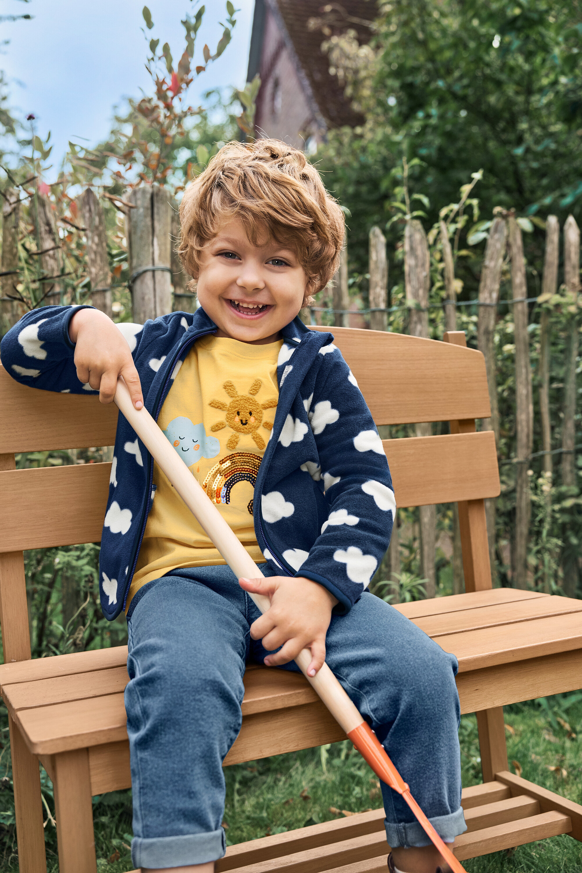 Uśmiechnięty chłopiec siedzi na małej ławce ogrodowej z półką i trzyma łopatę.