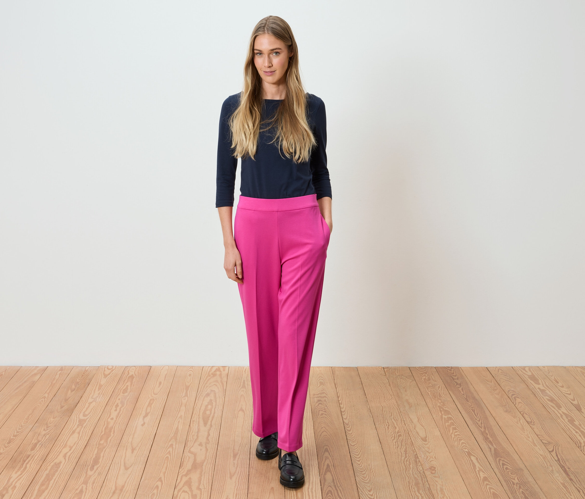 Kobieta stoi w granatowej bluzce i różowych spodniach na tle białej ściany.