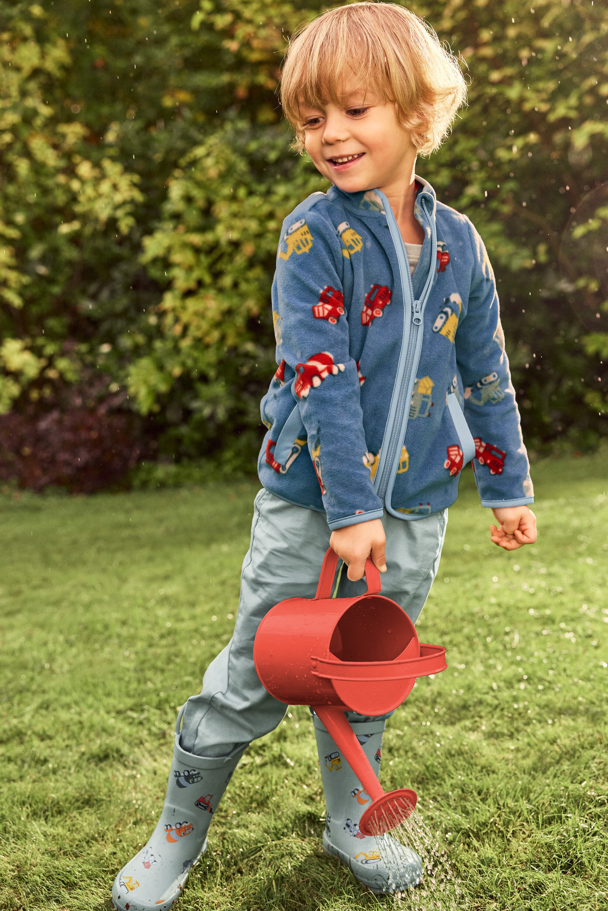 Uśmiechnięty blondwłosy chłopiec podlewa zielony trawnik czerwoną konewką. Ma na sobie niebieską kurtkę w pojazdy i szare kalosze również w pojazdy.