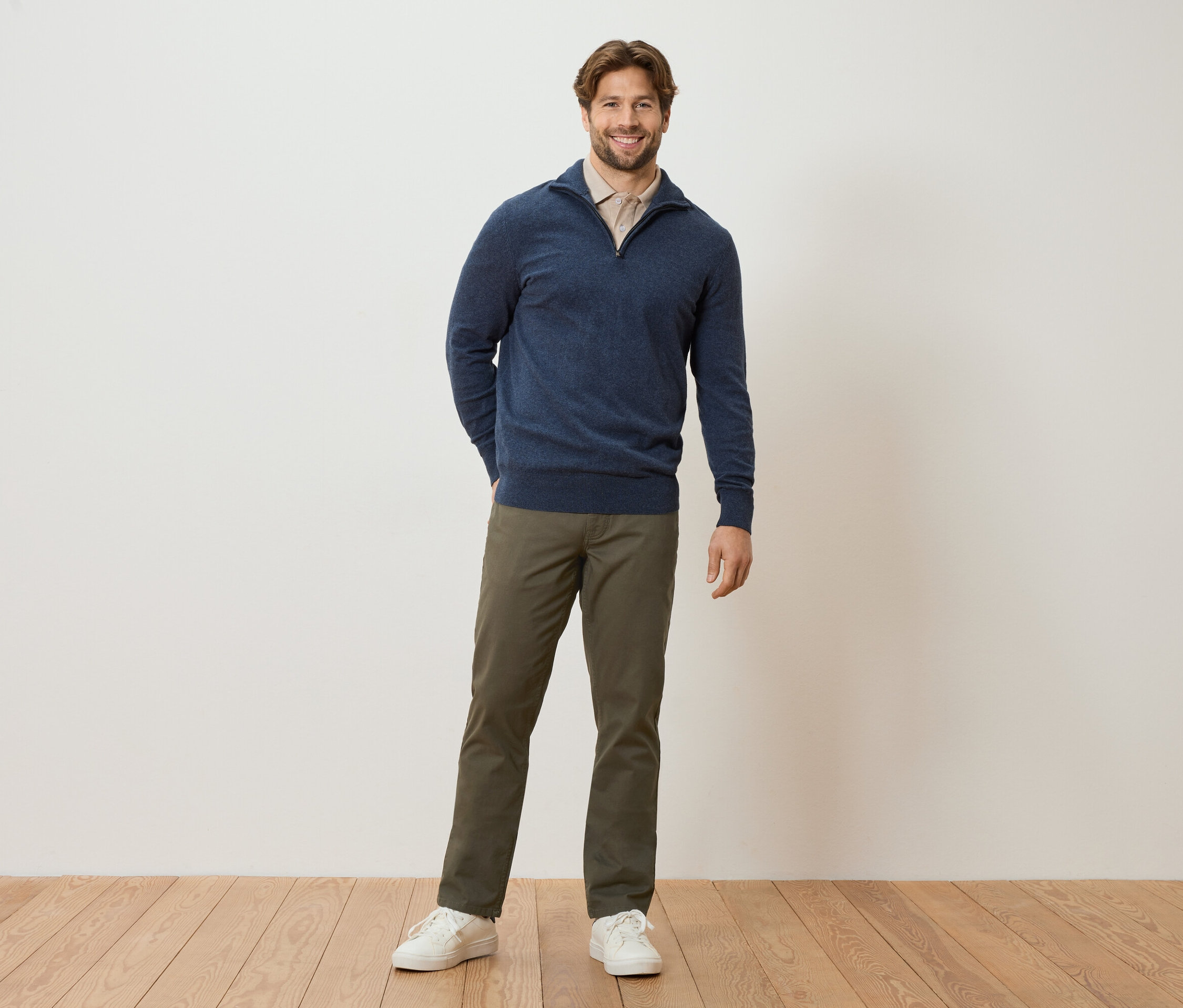 Mężczyzna stoi z rękami w kieszeniach na tle ściany. Ma na sobie granatowy sweter, jasnobrązową bluzkę, khaki spodnie i białe tenisówki.