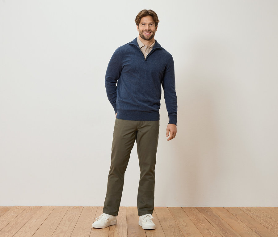 Mężczyzna stoi z rękami w kieszeniach na tle ściany. Ma na sobie granatowy sweter, jasnobrązową bluzkę, khaki spodnie i białe tenisówki.