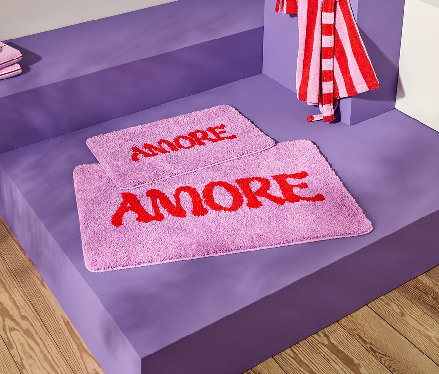 Zdjęcie dwóch lawendowych dywaników łazienkowych z czerwonym napisem "AMORE" na fioletowej powierzchni, wraz z ręcznikami w paski.