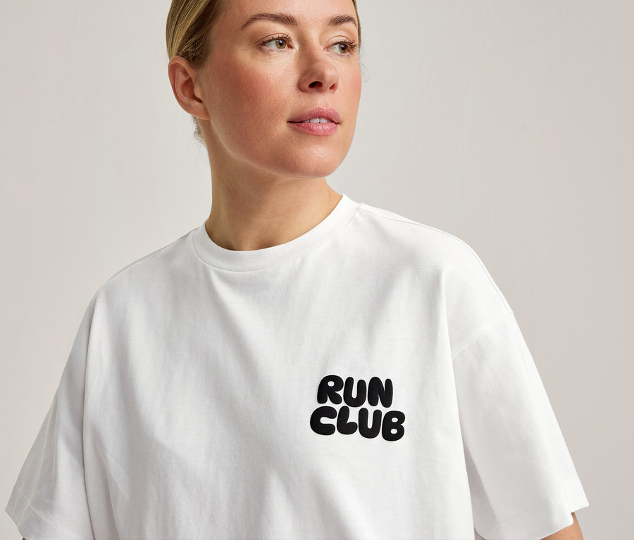 Kobieta ma na sobie białą koszulkę sportową z napisem „RUN CLUB” na piersi.