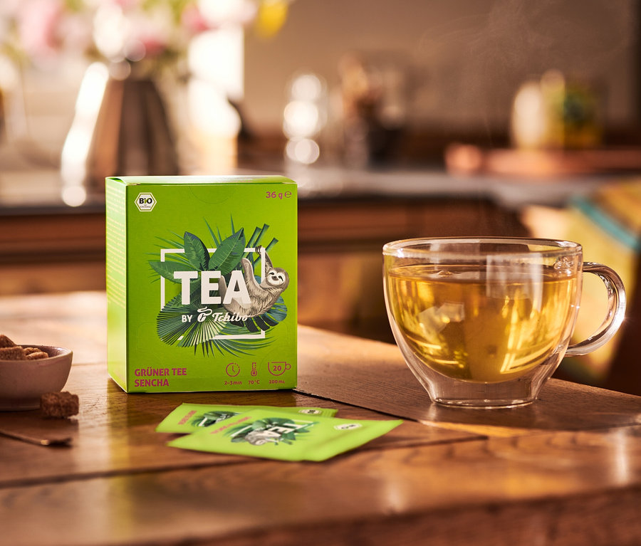 Na drewnianym stole stoi opakowanie BIO TEA by Tchibo Sencha 8x20x1,8 g, miska z cukrem i filiżanka herbaty.