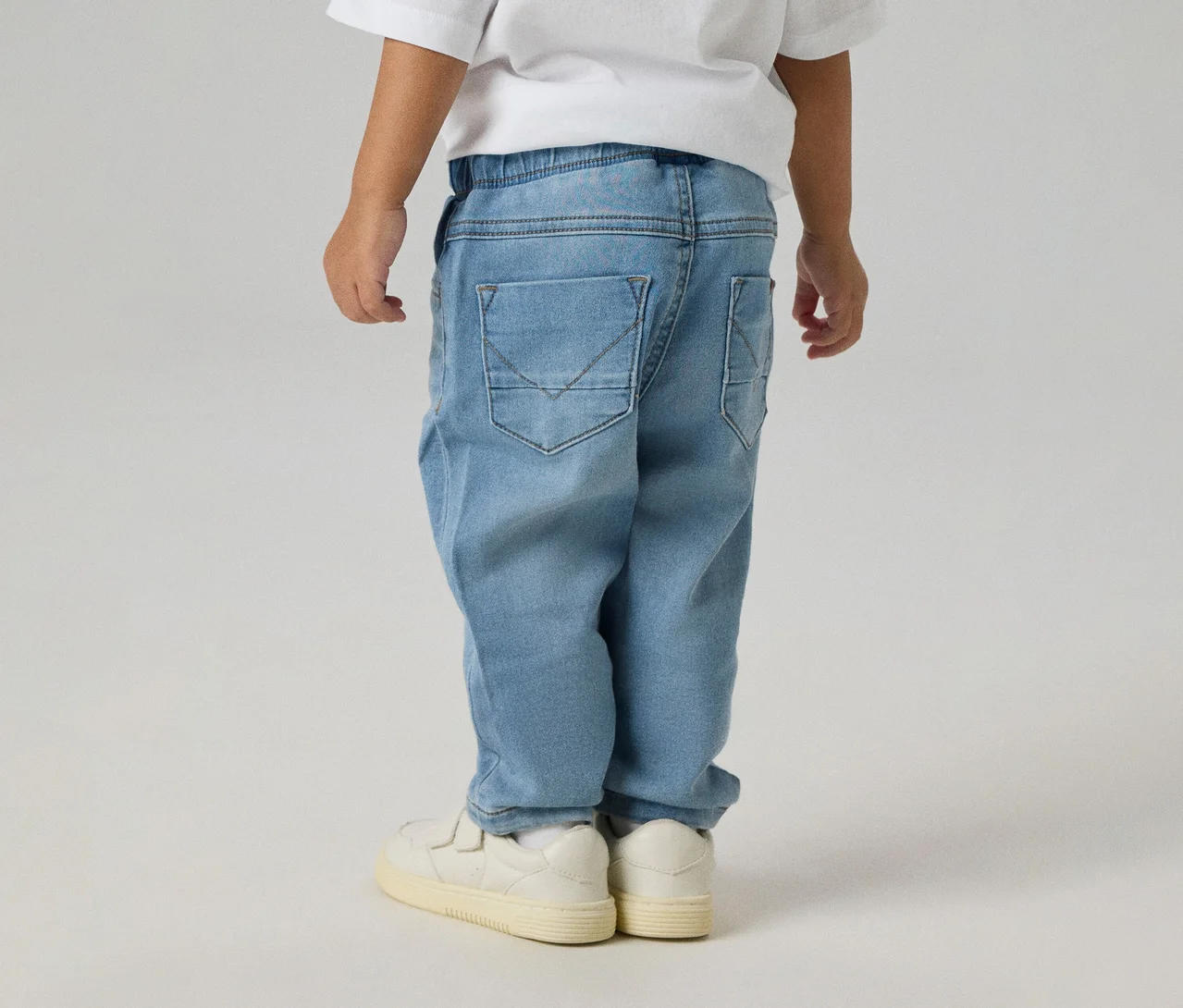 Dziecko od tyłu w jasnoniebieskich dżinsach NAME IT Slim Sweat Jeans z tylnymi kieszeniami i białych tenisówkach.
