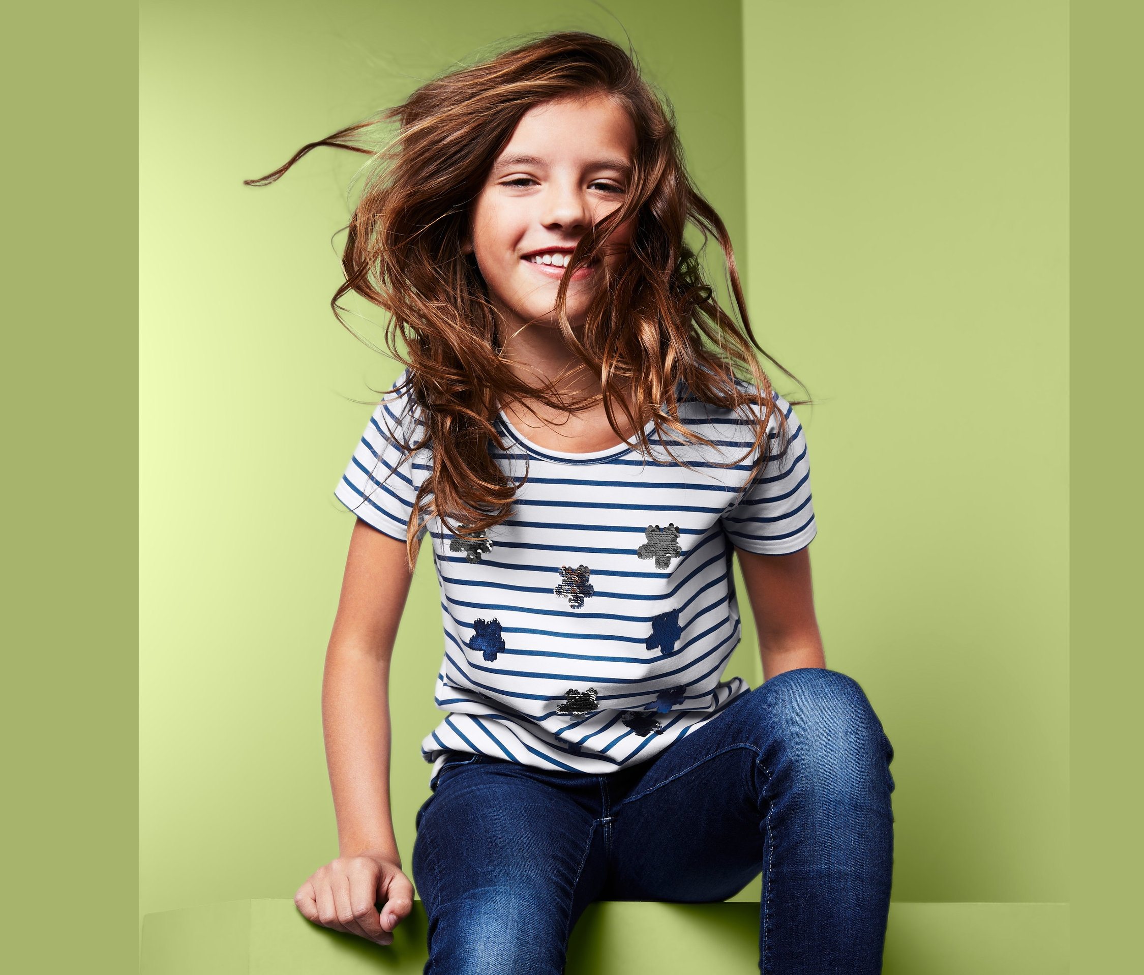 Dziewczyna siedzi na zielonej powierzchni, ubrana w koszulkę dziewczęcą z krótkim rękawem z dwustronnymi cekinami, dżinsy i trampki.