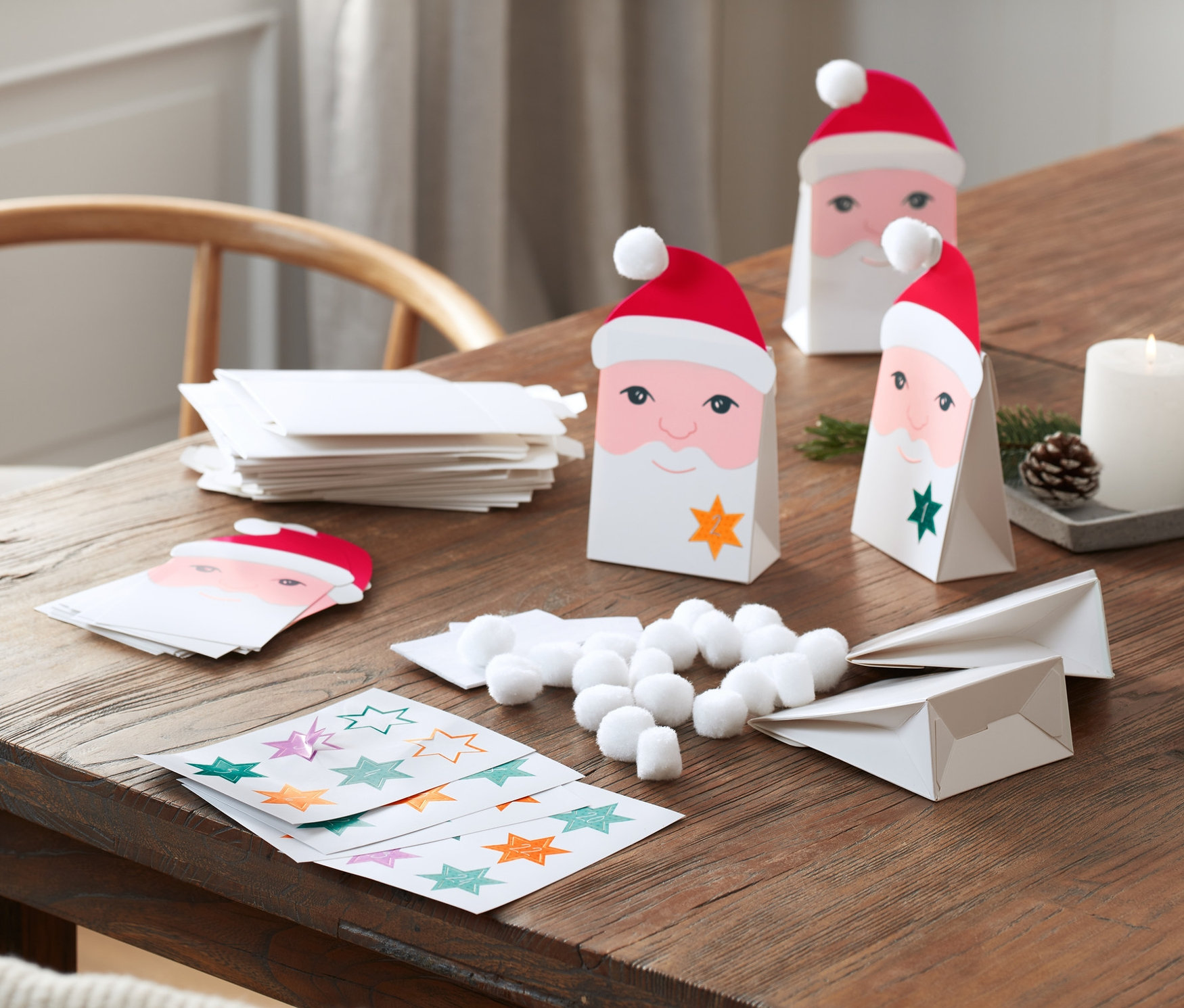 Na drewnianym stole leży zestaw do samodzielnego wykonania kalendarza adwentowego: papierowe torebki z buzią Świętego Mikołaja, naklejki z gwiazdami, kulki z waty i świeczki.