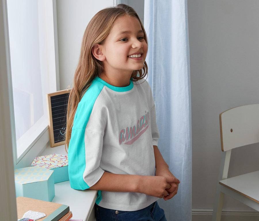 Uśmiechnięta dziewczyna opiera się o parapet, ubrana w koszulkę z bawełny ekologicznej z turkusowymi rękawami.