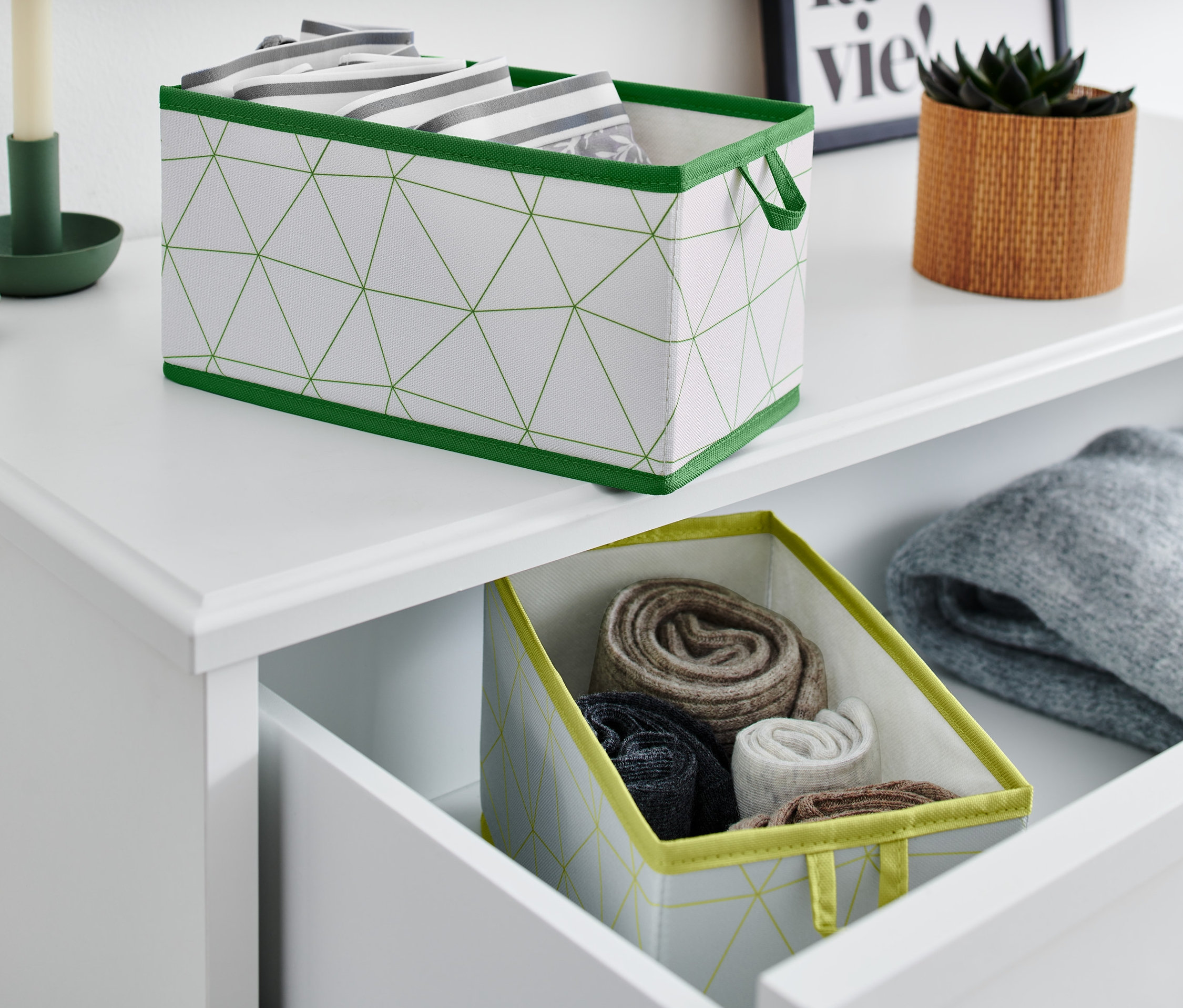 Dwa średnie organizery do szuflad, jeden na górze komody, a drugi w otwartej szufladzie ze zwiniętymi ubraniami.