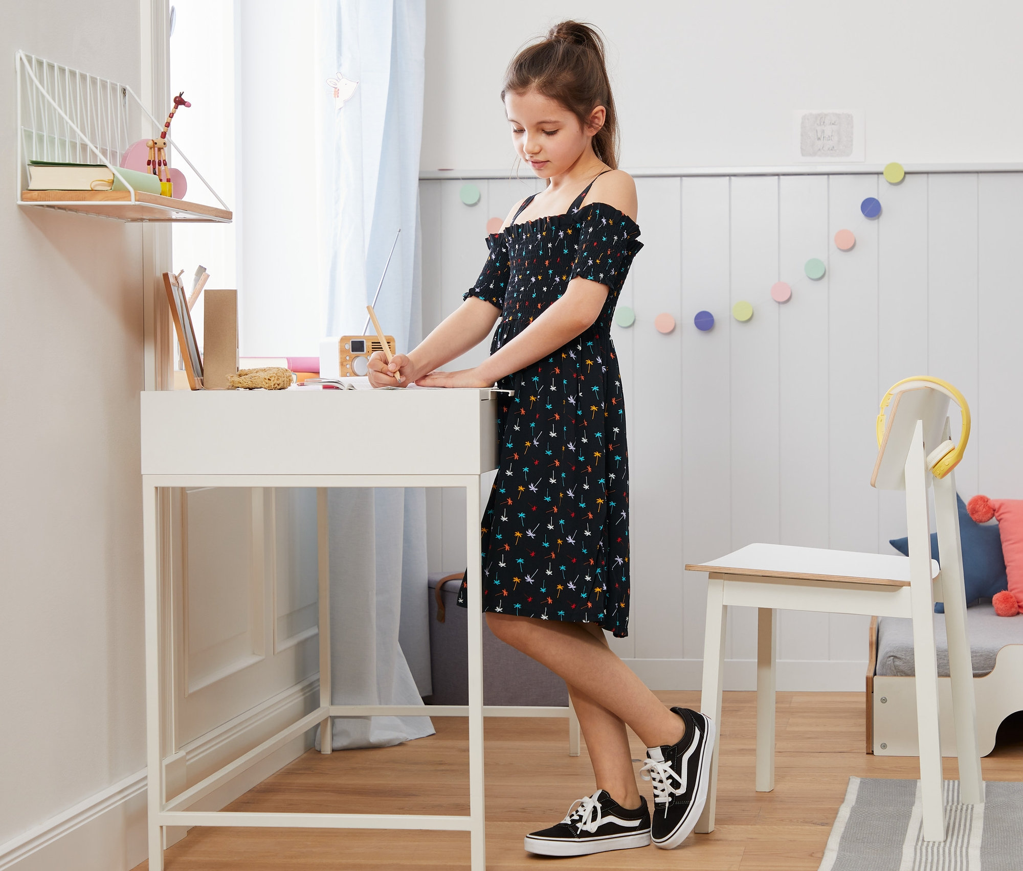 Dziewczyna pisze przy białym biurku w sukience z odkrytymi ramionami.