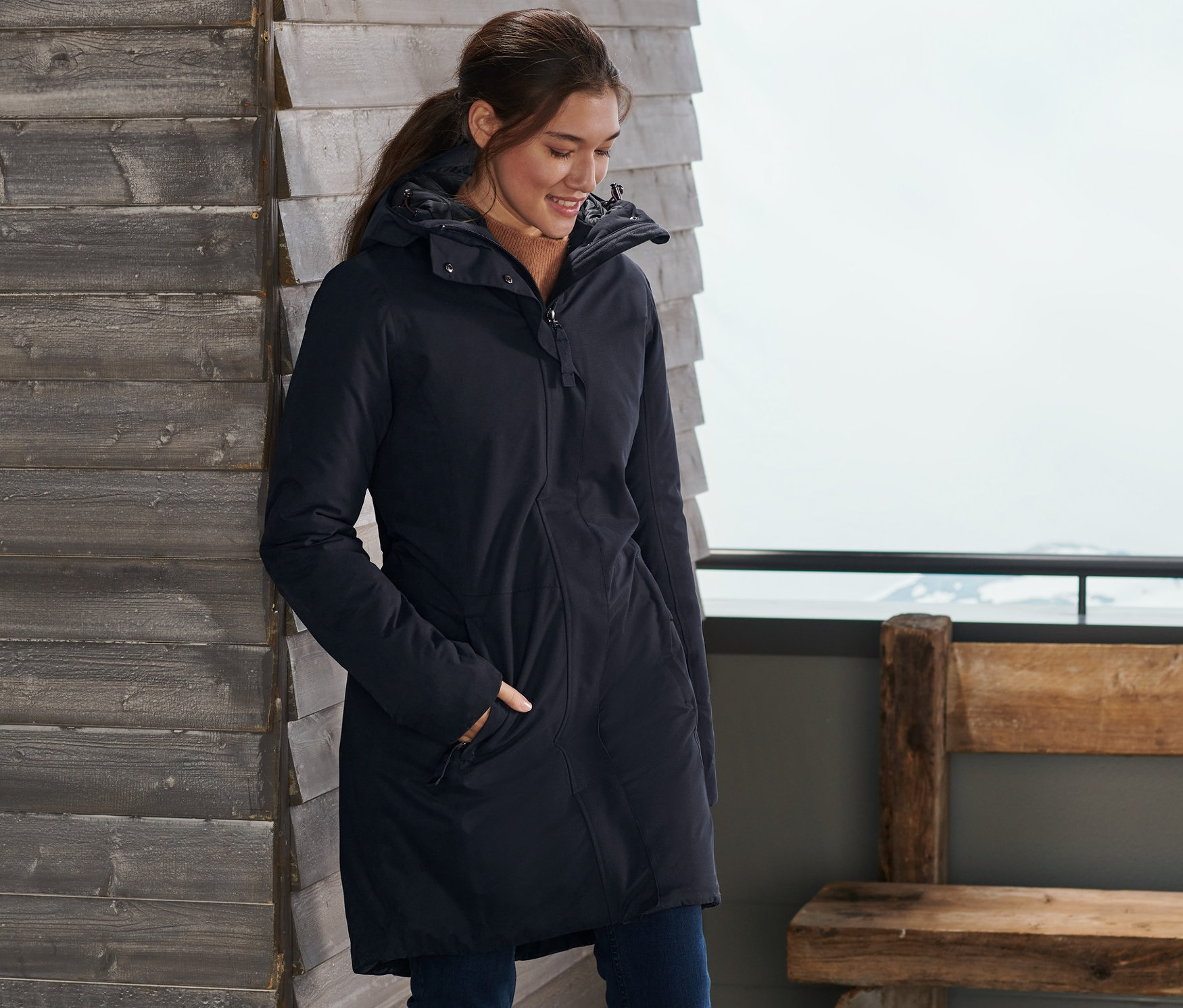 Kobieta stoi przy drewnianej ścianie z nartami, ubrana w niebieski płaszcz ciepłochronny.