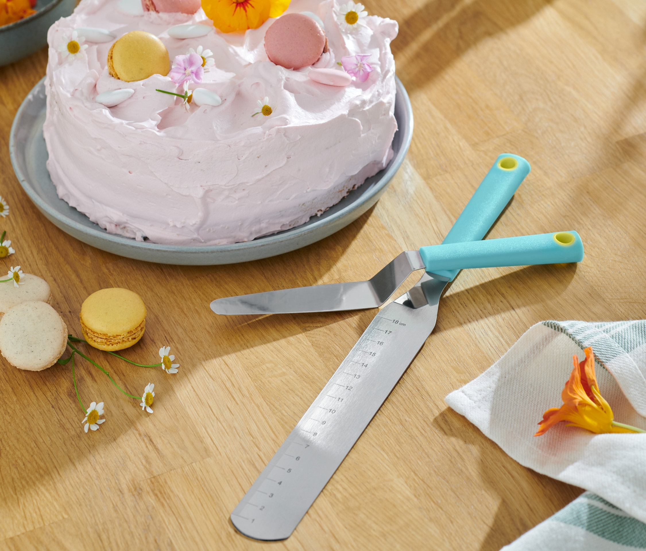 Dwie krzyżujące się Szpatułki do ciast, 2 sztuki leżą na drewnianym stole obok ciasta z różowym lukrem i makaronikami.