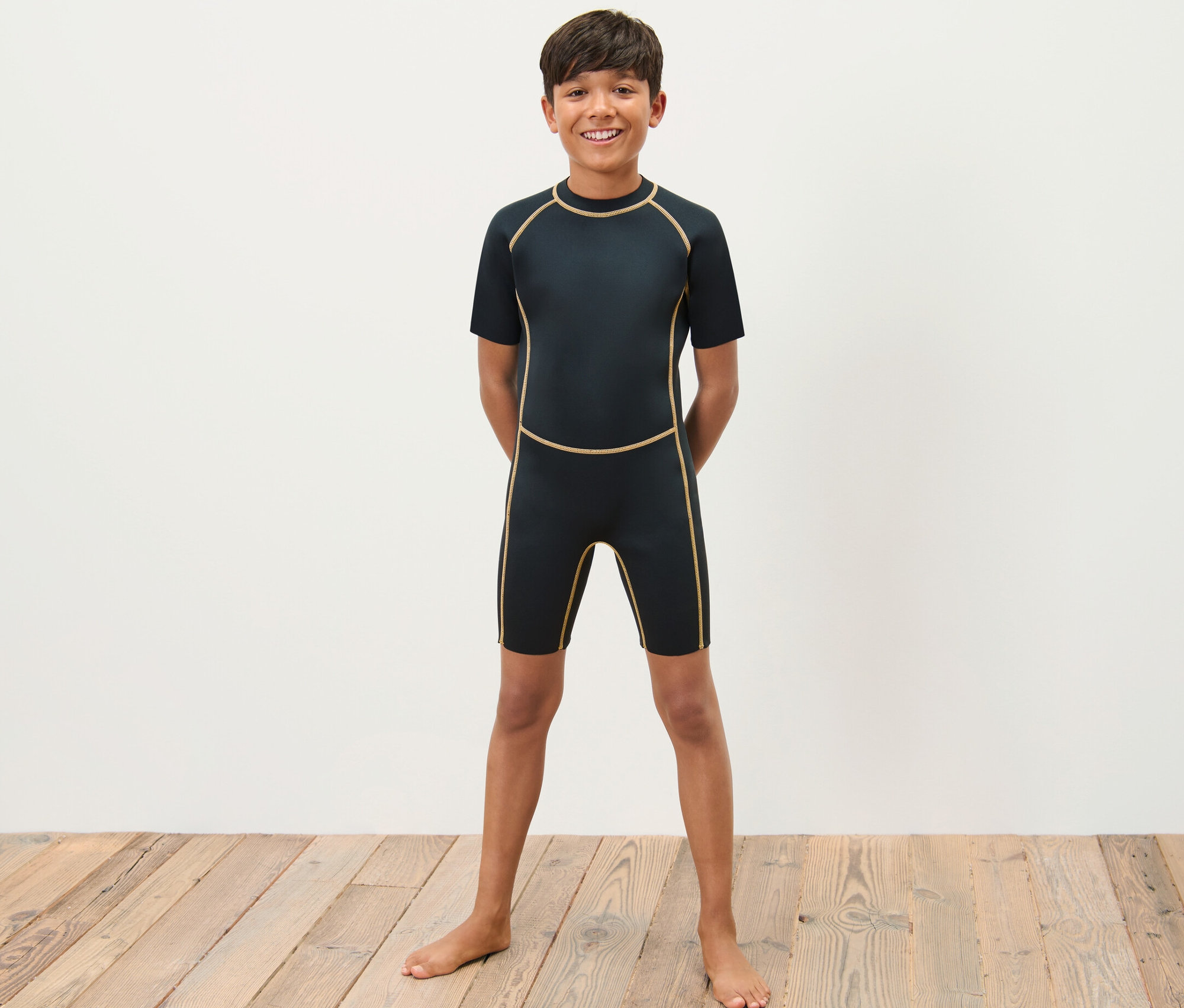 Chłopiec w dziecięcym kombinezonie piankowym stoi boso na drewnianej podłodze przed białą ścianą.