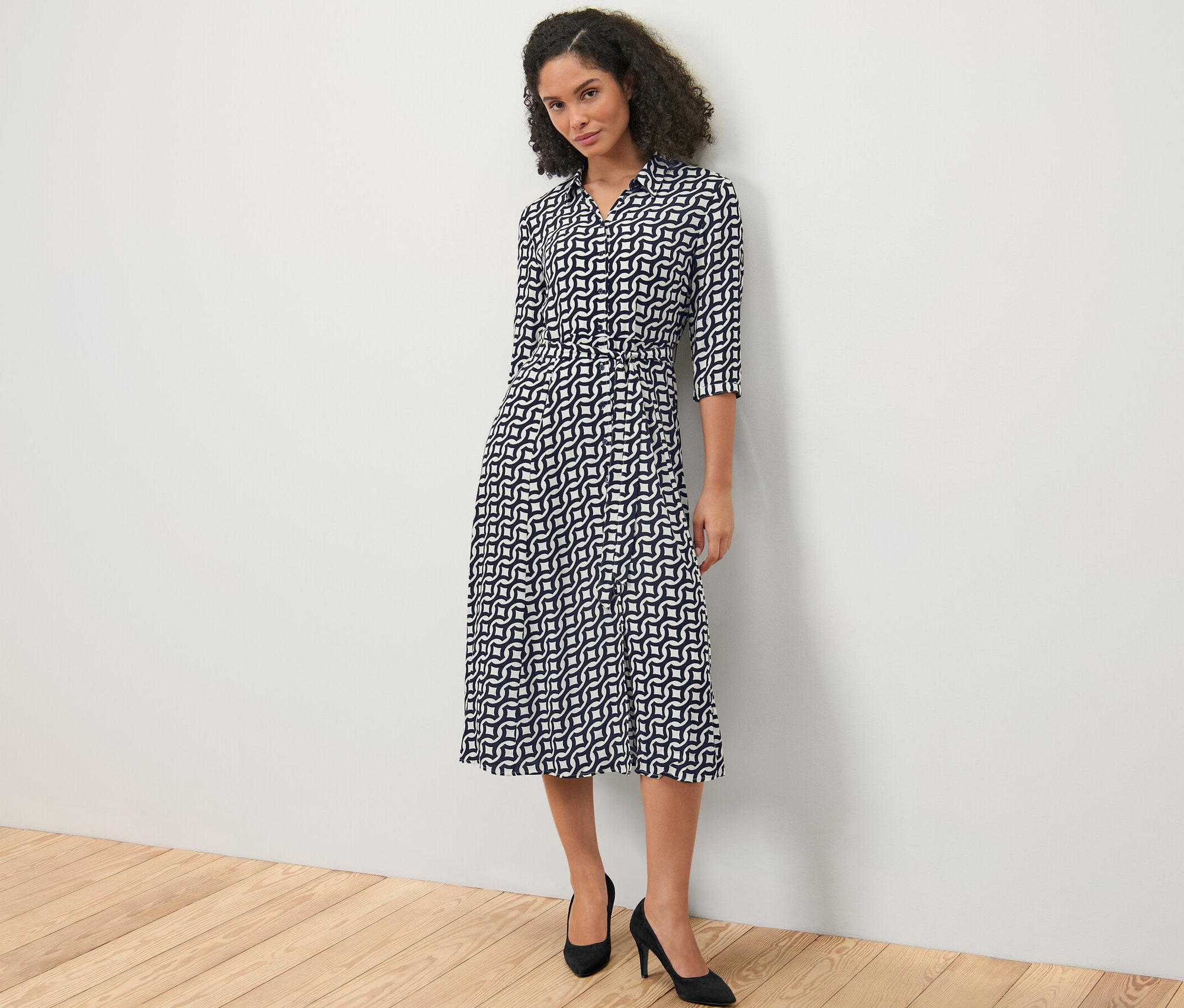 Kobieta stojąca na tle białej ściany ma na sobie wzorzystą sukienkę koszulową i czarne buty na obcasie.