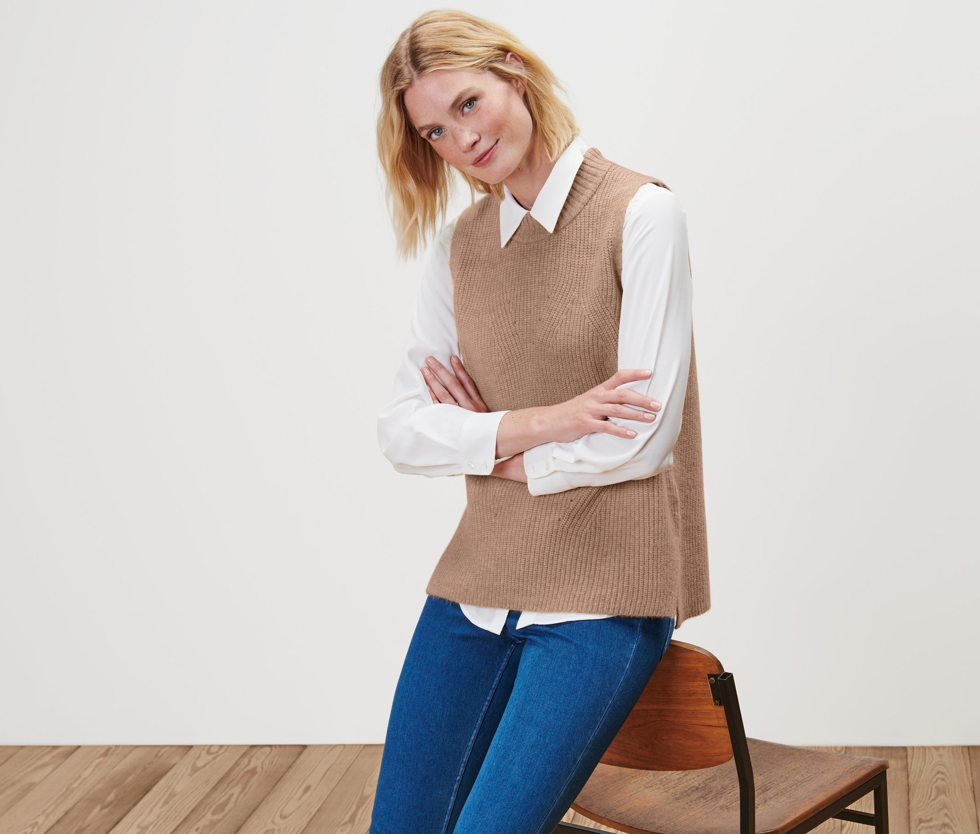 Blondwłosa kobieta pozuje na krześle w beżowym melanżowym bezrękawniku dzianinowym, białej koszuli i niebieskich dżinsach.