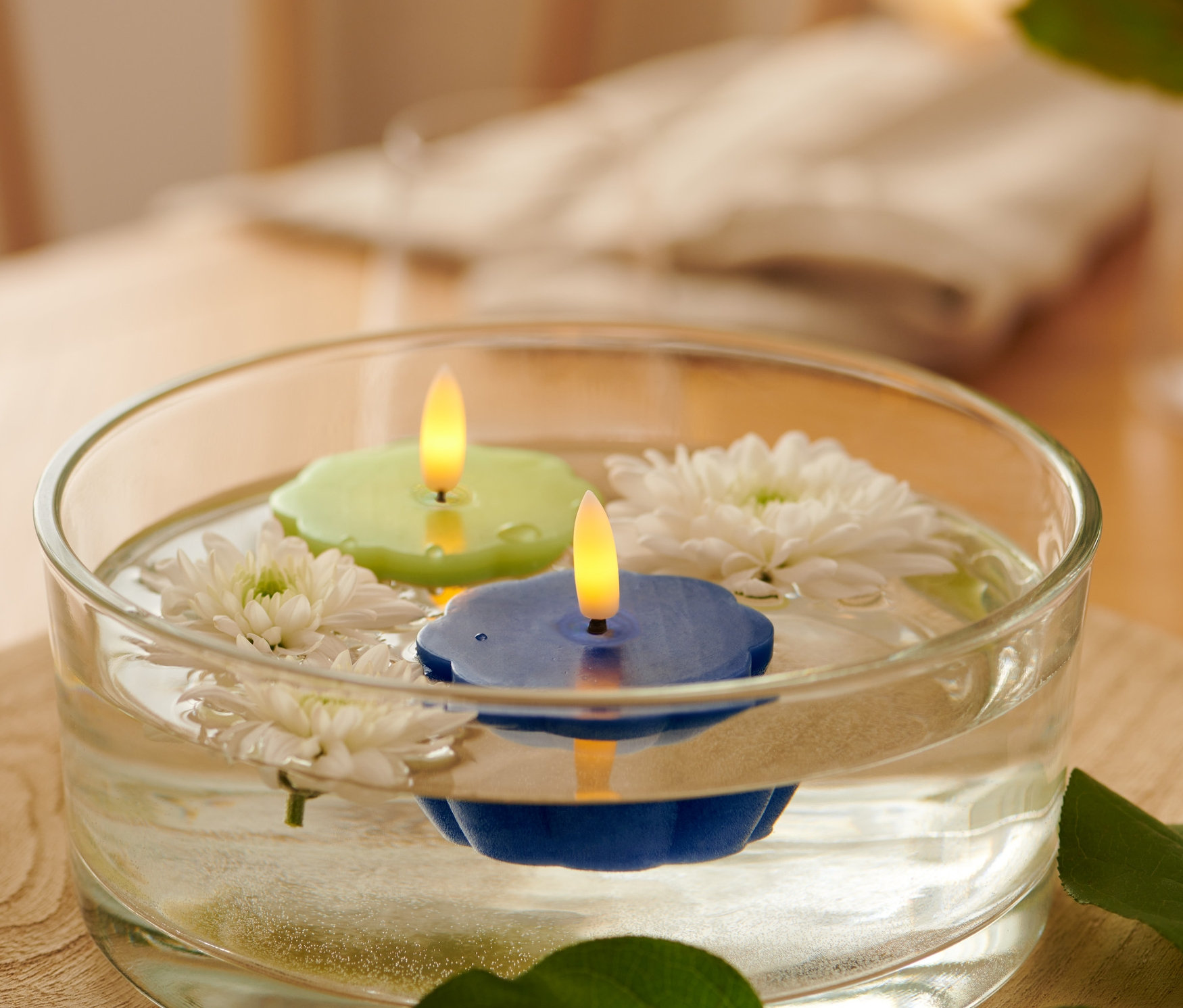 W szklanej misce pływają dwie świeczki LED typu tea light z prawdziwego wosku, zielona i niebieska, oraz białe kwiaty.