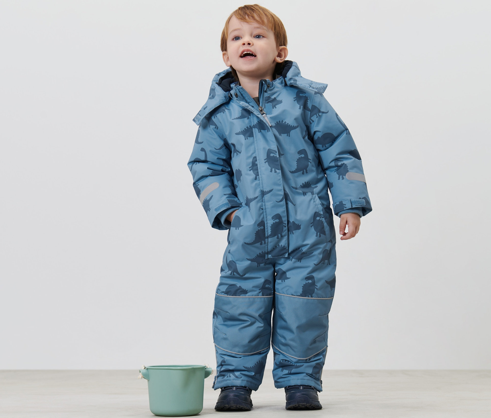 Chłopiec stoi w dziecięcym kombinezonie zimowym w dinozaury i patrzy w górę. Obok niego stoi zielone wiaderko.