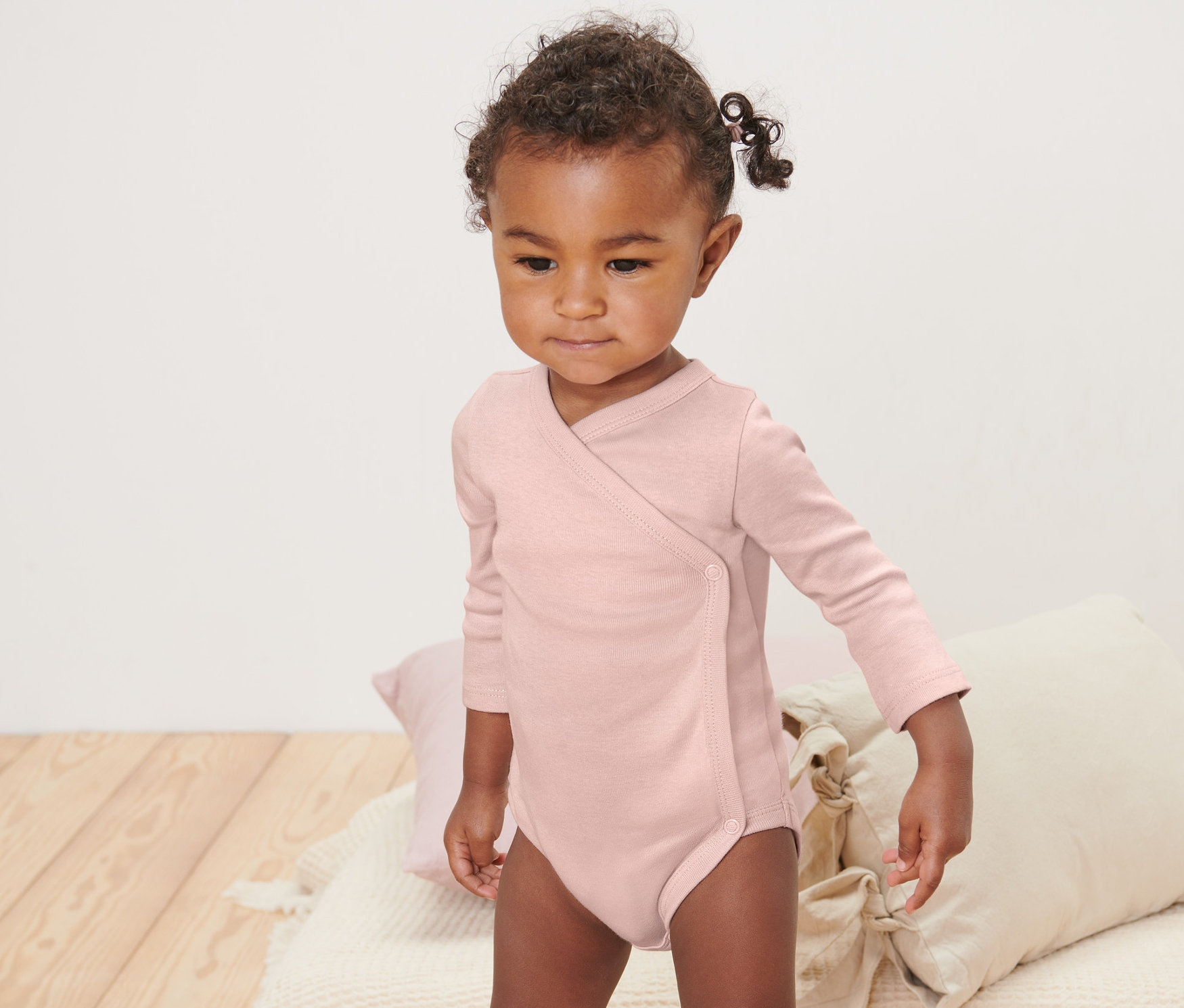 Małe dziecko w różowym body z długim rękawem stoi na kocu. Na kocu leżą poduszki. Body niemowlęce rosnące z dzieckiem, 5 par, różowe.