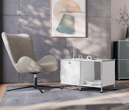 W salonie stoi fotel obrotowy obok białego metalowego stolika „CN3” z jedną klapą i kółkami. W tle znajduje się szara metalowa szafka typu sideboard „CN3” z 2 klapami oraz chromowana metalowa lampa stołowa.
