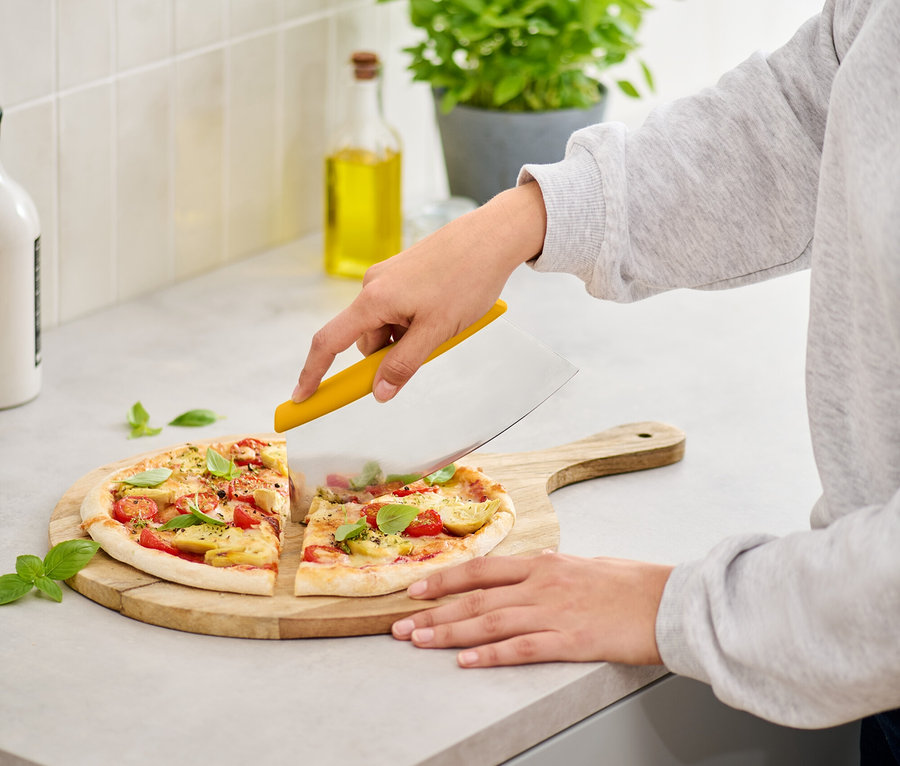 Kobieta kroi pizzę z pomidorami i bazylią na drewnianej desce do krojenia żółtym krajalnikiem do pizzy.