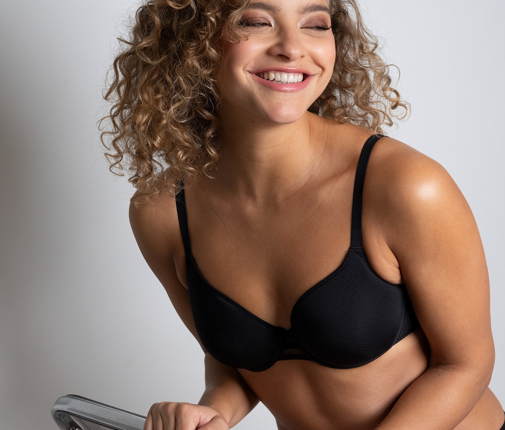 Uśmiechnięta kobieta siedzi na przezroczystym krześle, ubrana w czarny biustonosz typu spacer Passionata „Nais”.