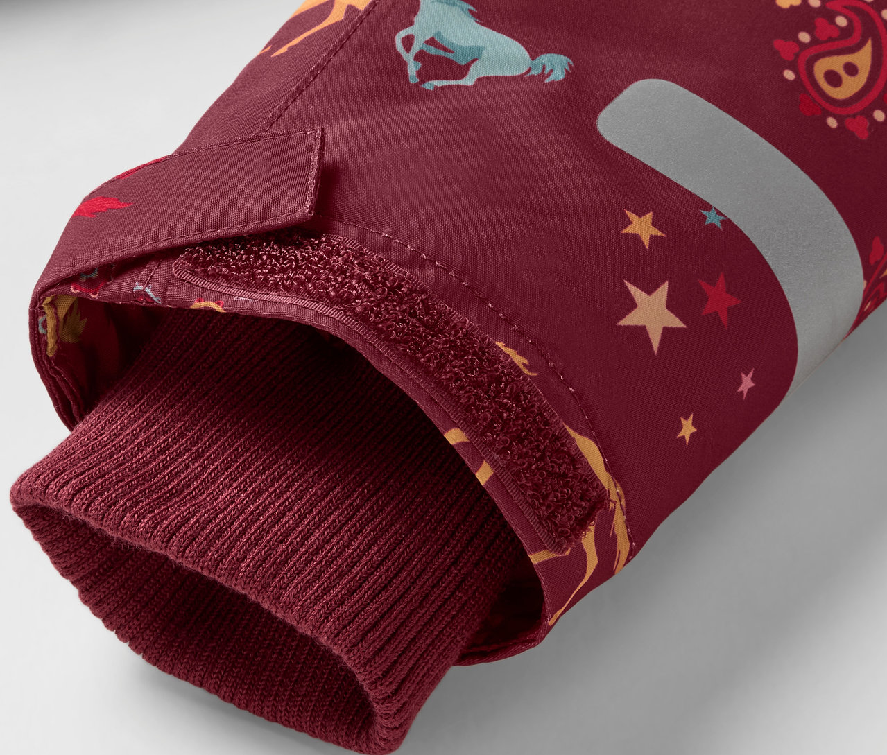 Detal bordowego rękawa kurtki z nadrukiem koni i gwiazd, wykończonego bordowym ściągaczem.