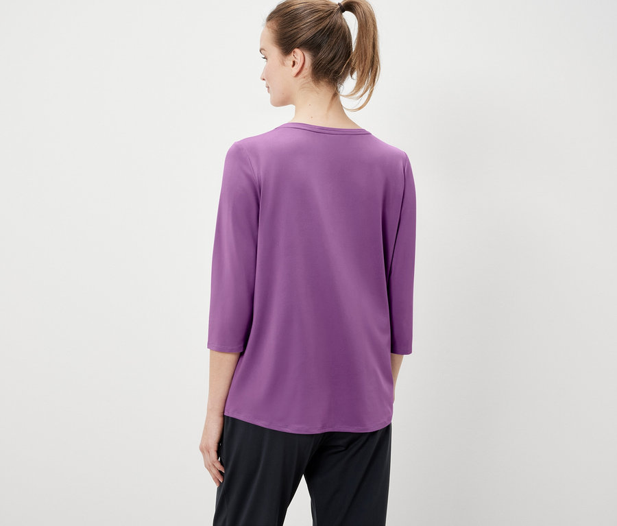 Z tyłu widać kobietę z włosami związanymi w koński ogon, ubraną w fioletową koszulkę z rękawami trzy czwarte i czarne spodnie.