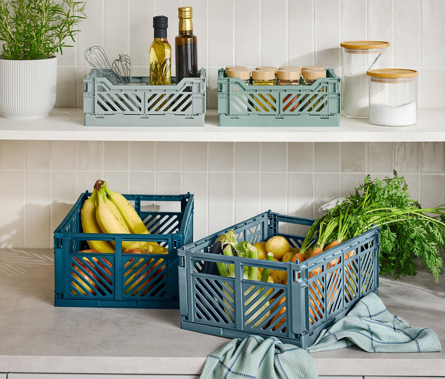 Na blacie kuchennym znajdują się trzy składane pojemniki do przechowywania, jeden niebieski z bananami, jeden szary z warzywami i jeden szary z przyborami kuchennymi.