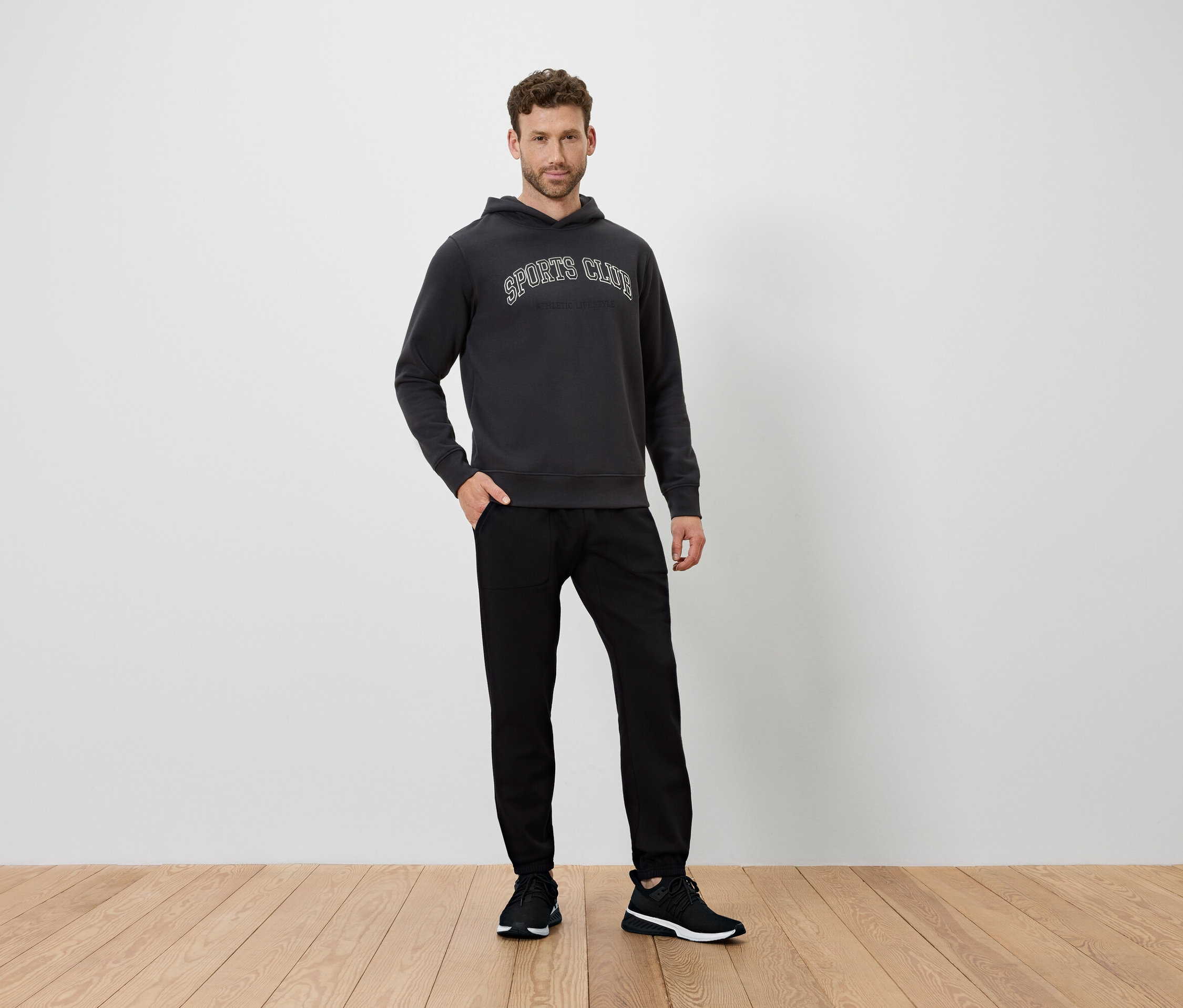 Mężczyzna stoi na drewnianej podłodze, ubrany w ciemnoszarą bluzę z kapturem i czarne spodnie.
