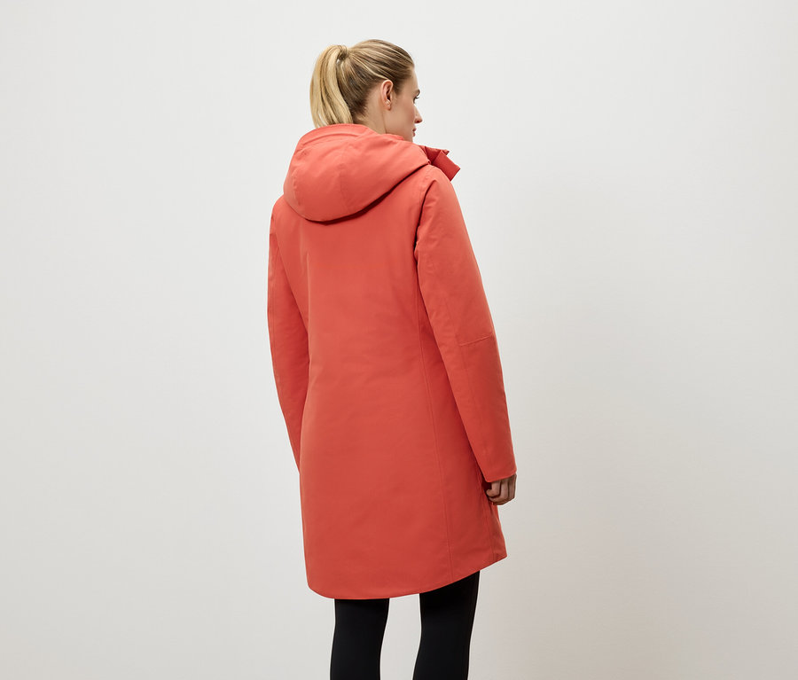 Kobieta tyłem do kamery ma na sobie koralowy płaszcz z kapturem i czarne legginsy.