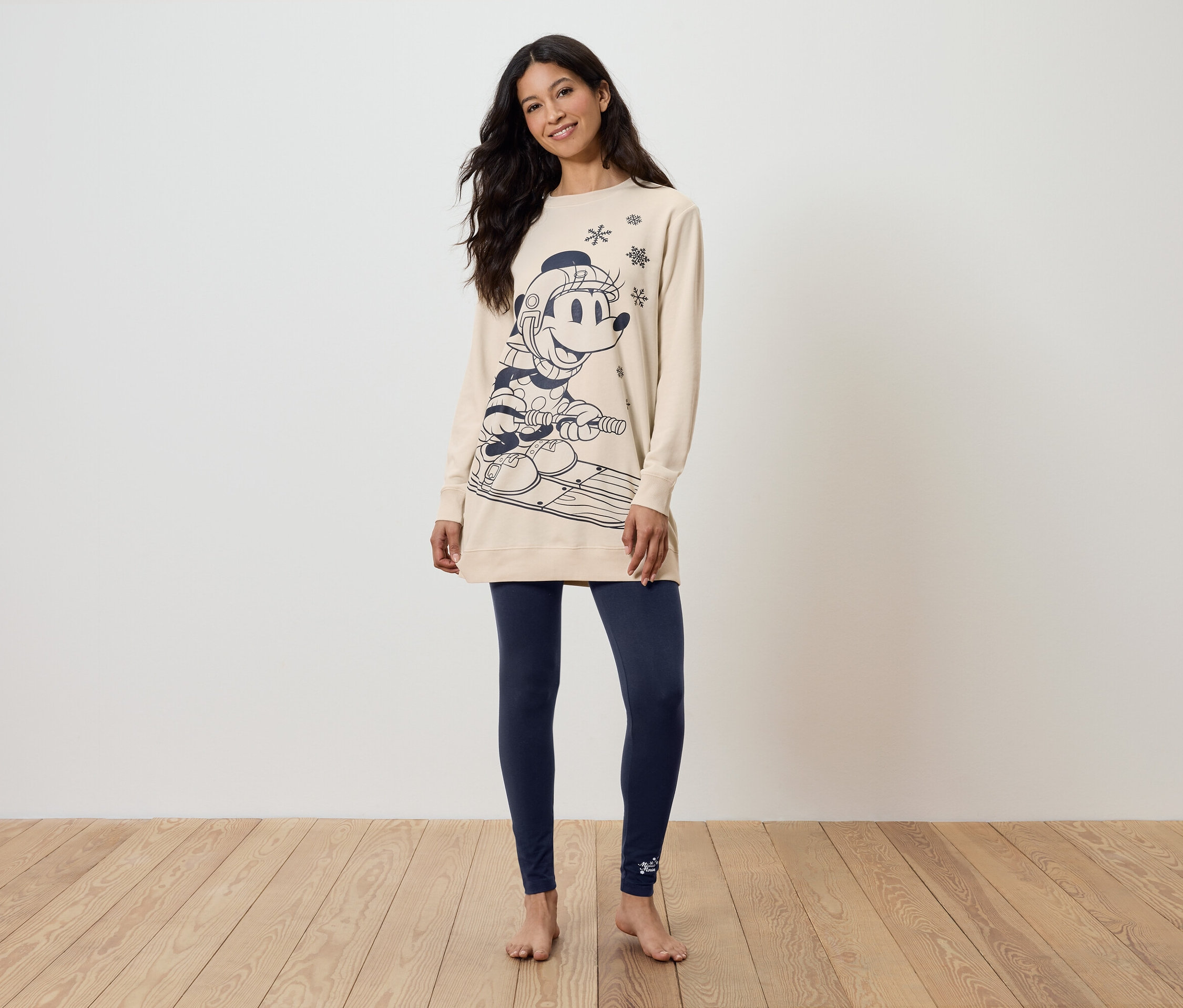 Kobieta stoi boso, ubrana w długą, beżową bluzę z nadrukiem i granatowe legginsy.