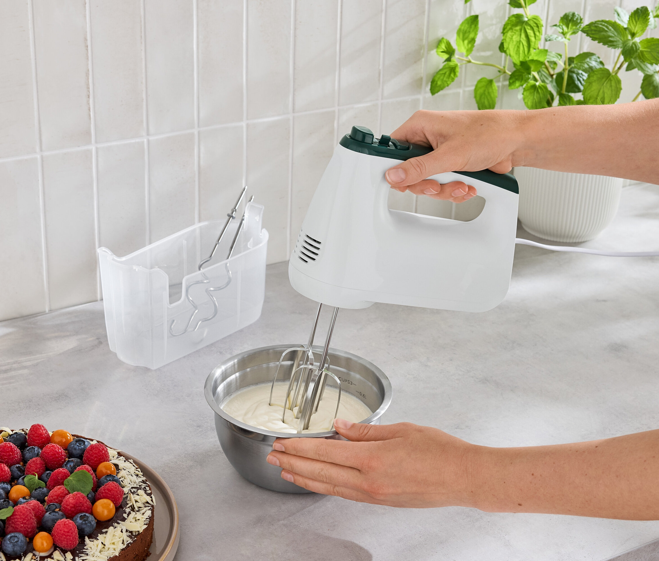 Kobieta miesza ciasto w misce mikserem ręcznym obok ciasta z owocami.