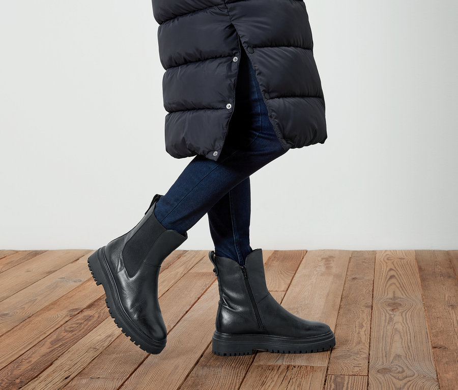 Nogi osoby w dżinsach i czarnych skórzanych butach na drewnianej podłodze. Osoba ma na sobie długą, czarną kurtkę.