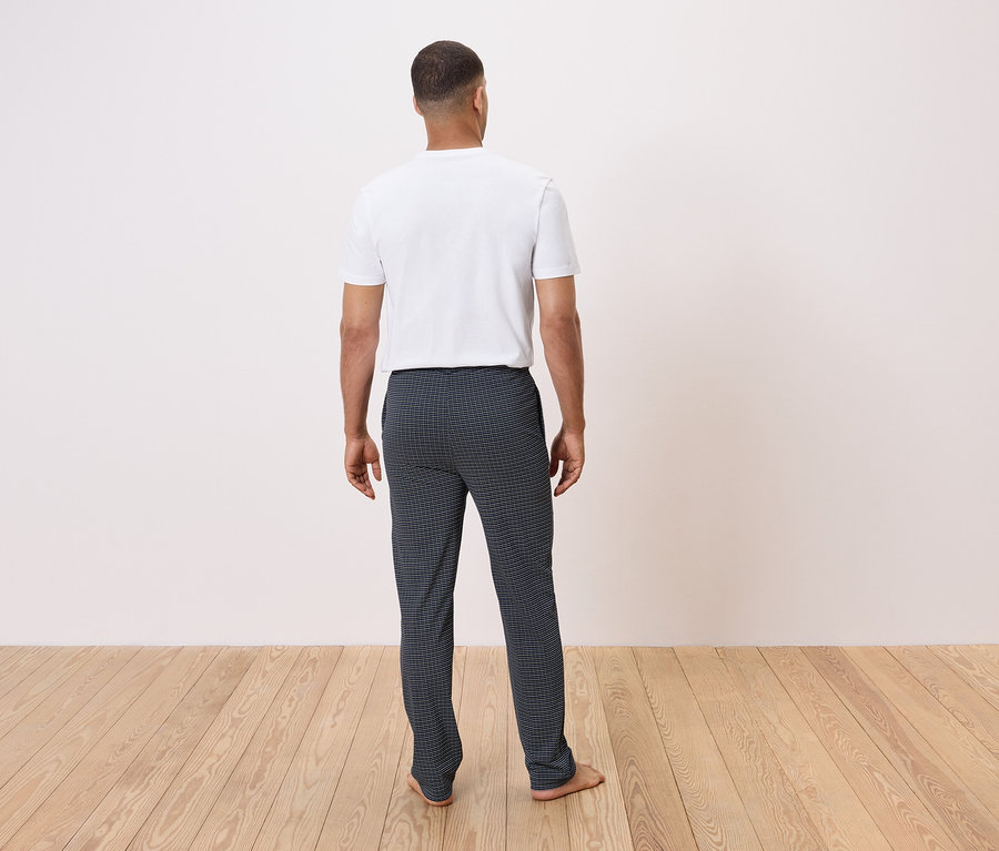 Mężczyzna stoi tyłem do kamery na drewnianej podłodze, ubrany w białą koszulkę i ciemne spodnie w kratę.