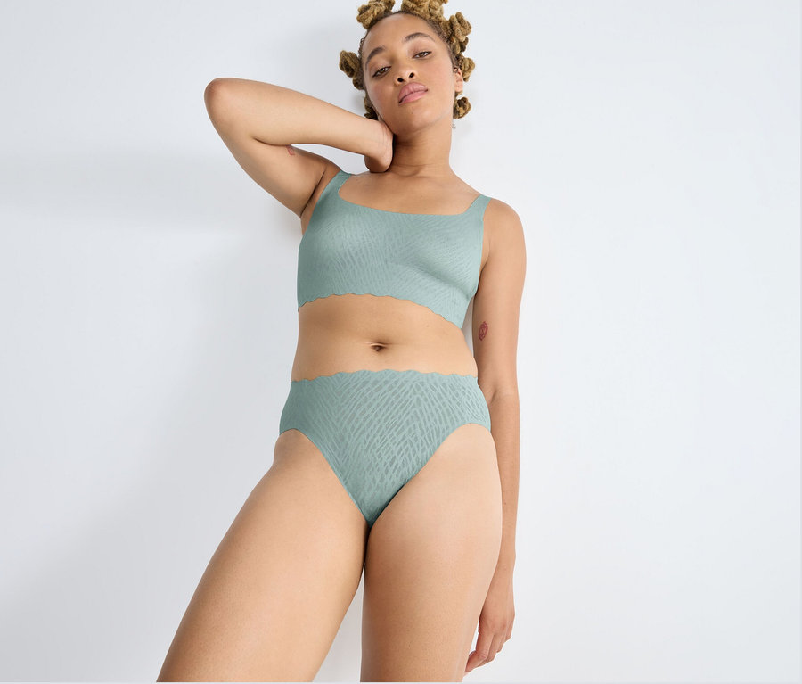 Kobieta z blond dredami stoi przy białej ścianie, ubrana w figi high leg sloggi ZERO Feel Bliss w kolorze misty turquoise i pasujący top. Ma uniesioną rękę za głową i patrzy w kamerę. Oba elementy mają delikatny wzór i falowane krawędzie.