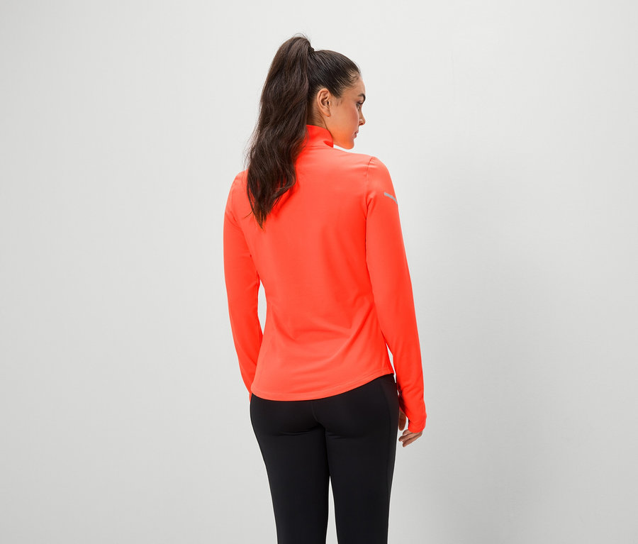 Kobieta tyłem do kamery ma na sobie pomarańczową bluzkę z długim rękawem i czarne legginsy.