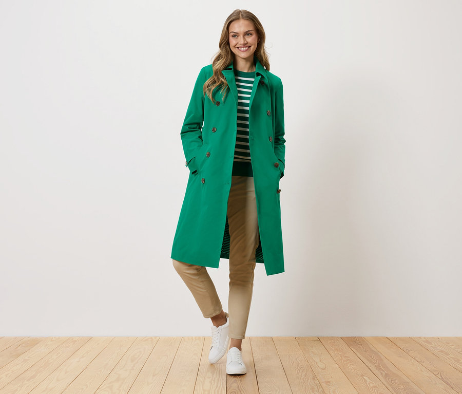 Kobieta w zielonym płaszczu, w paski sweter, beżowe spodnie i białe trampki stoi na tle białej ściany.