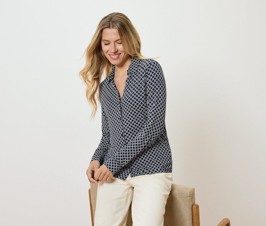 Kobieta z długimi blond włosami w wzorzystej bluzce i jasnych spodniach siedzi na krześle i uśmiecha się.